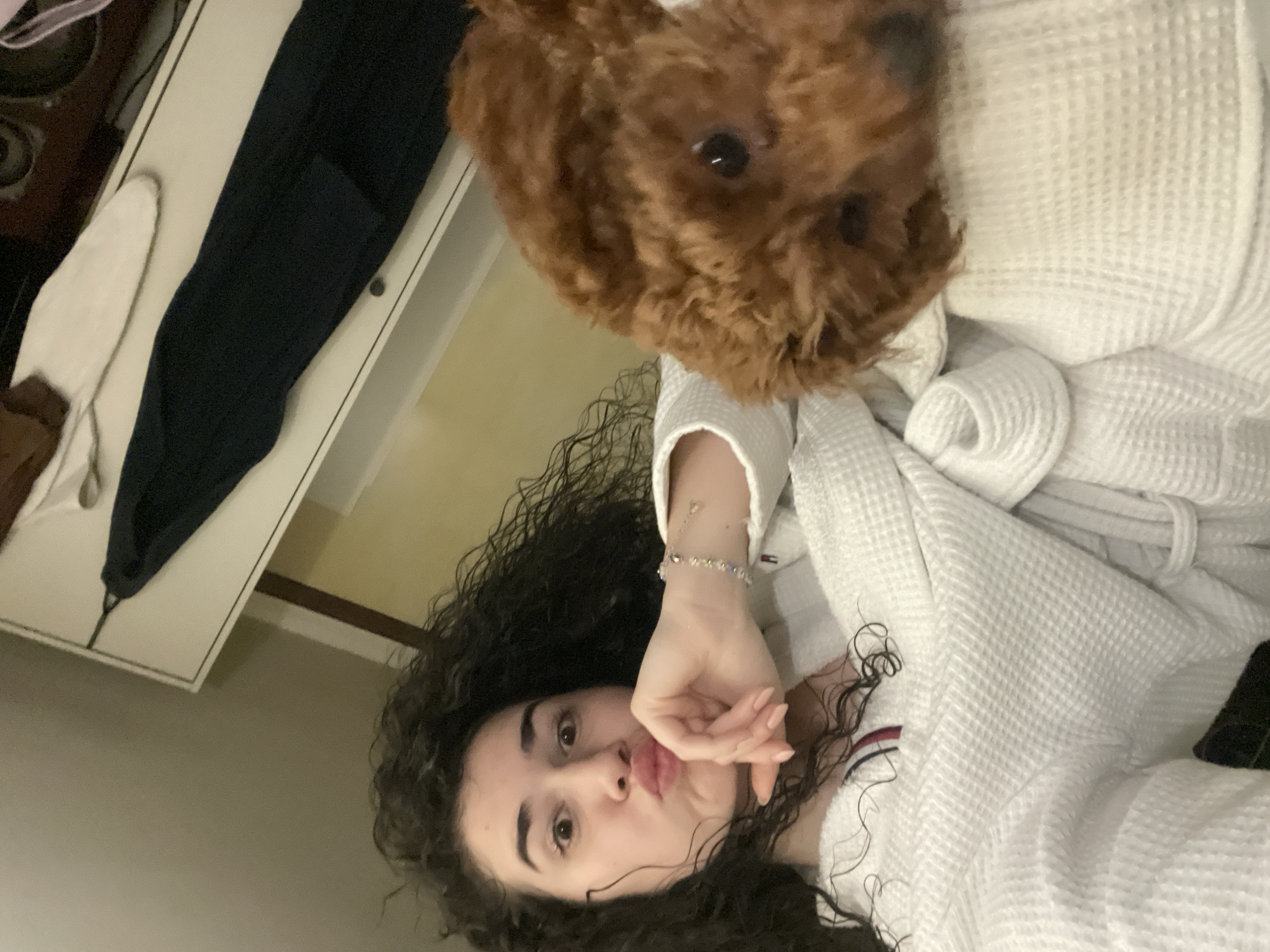 The image shows a person with curly dark hair, making a pouting face while lying down, wearing a white robe. A small brown poodle is positioned near the person's head, also partially visible. In the background, there's a white dresser with dark-colored clothing placed on it. The location is not discernible, therefore it's not possible to determine if it's in Sofia, Bulgaria.