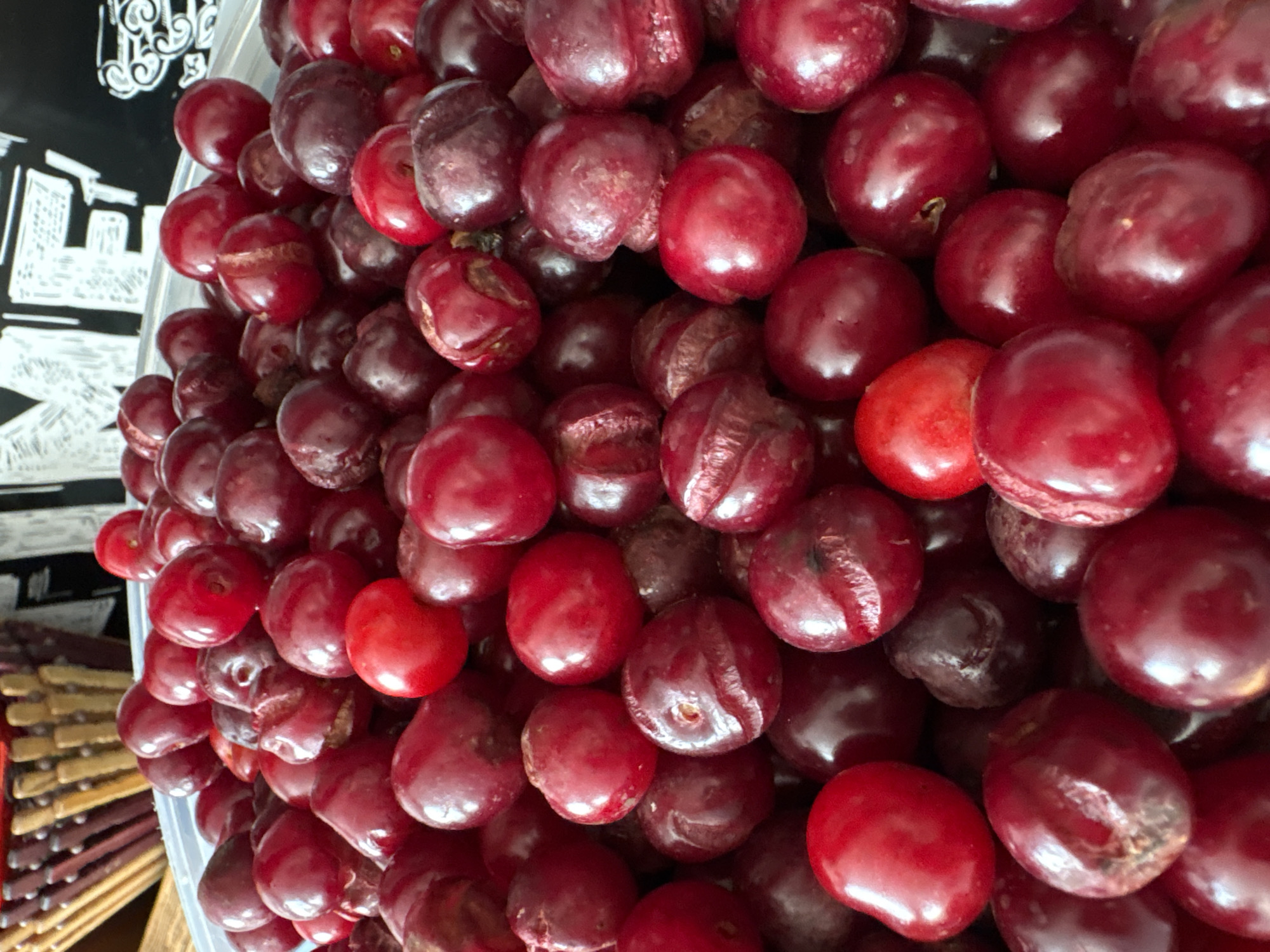 A close-up, overhead shot shows a large pile of ripe cherries. The cherries are a deep red color and appear to be fresh. Some of the cherries have stems, while others do not. The pile of cherries is so dense that it is difficult to distinguish individual fruits in some areas. The background is blurred, but appears to be a white surface with some writing on it. The lighting is bright and even, with no harsh shadows.