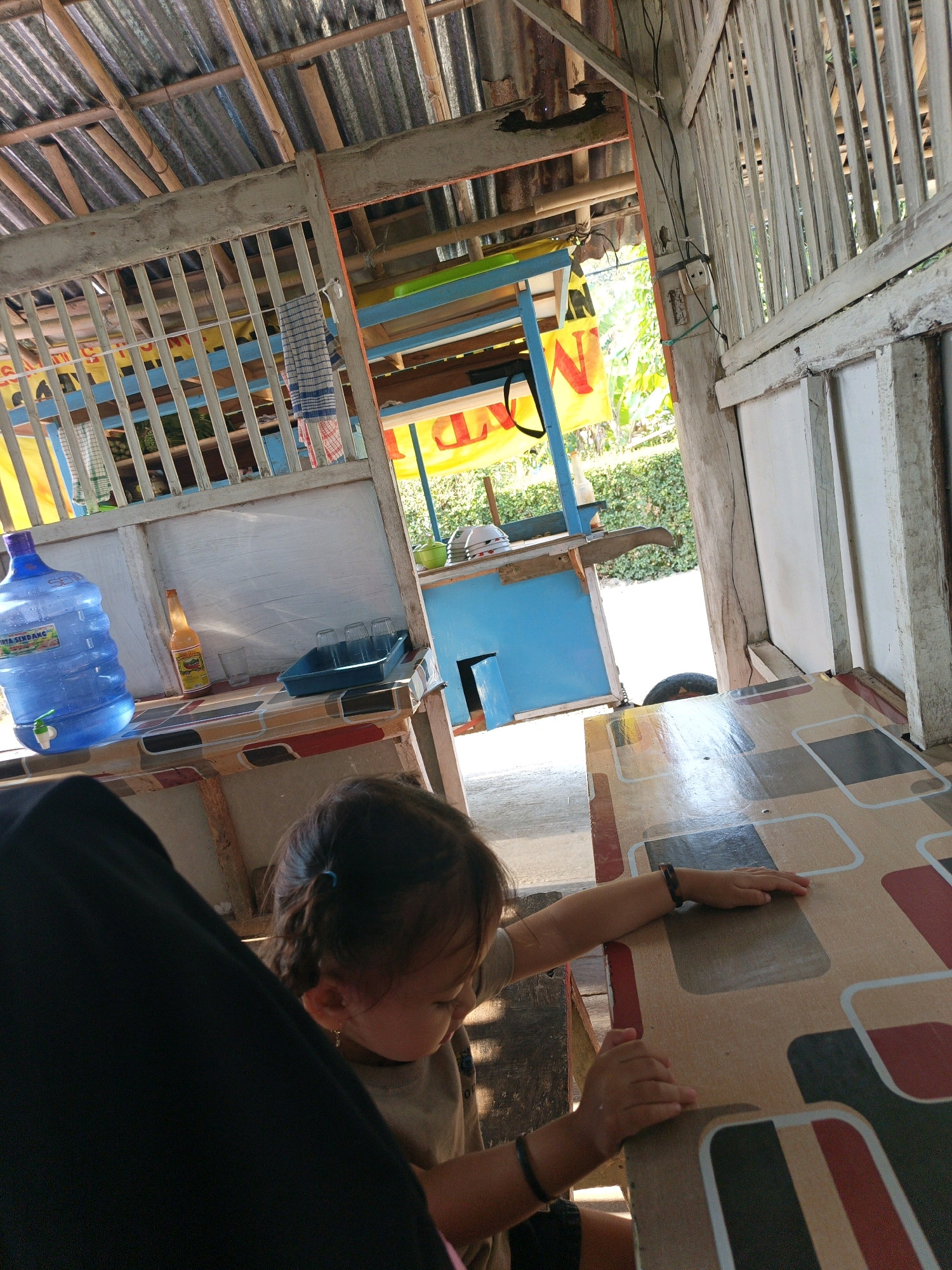 The image depicts a young child with dark hair, wearing a light-colored t-shirt and black shorts, sitting at a patterned table. The child's hands are resting on the table, and they are wearing bracelets on both wrists. A large blue water dispenser is visible on a counter to the left, along with a bottle of orange liquid, several glasses, and a blue tray. In the background, a blue cart is visible, and beyond that, lush green foliage and a yellow sign with red lettering. The setting appears to be an outdoor eatery or stall, likely in Kamulyan, Indonesia, characterized by its rustic wooden structure with a corrugated metal roof and bamboo supports. The overall scene is candid and appears to capture a moment of informal dining or waiting.