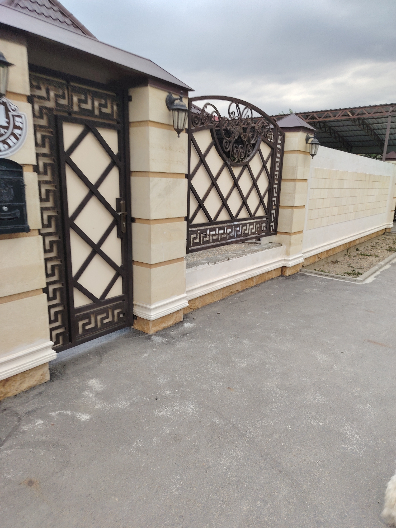 The image shows a stone fence and gate in the city of Volnoye, Russia. The fence is made of light-colored stone blocks and has a decorative metal gate. There are two black outdoor lamps attached to the fence posts. A dark-colored mailbox is mounted on the left side of the gate. The ground in front of the fence is paved with asphalt. The sky is overcast.