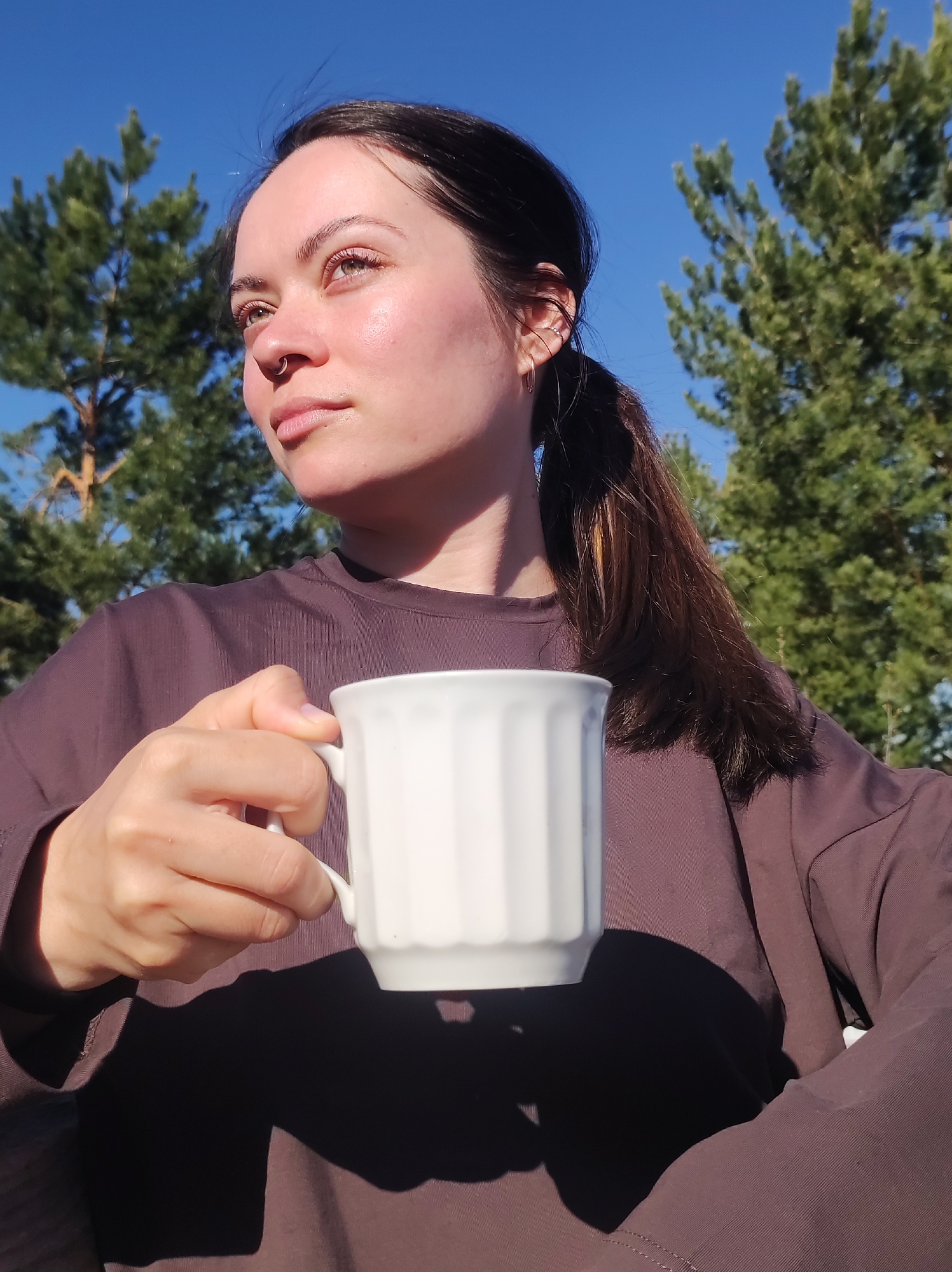 A woman with dark hair tied back in a ponytail holds a white mug outdoors. She is fair-skinned, with a nose ring, and wears a brown long-sleeved shirt. She looks off to the left. Evergreen trees and a clear blue sky are visible in the background. The image does not contain any information about the city of Фоки, Russia.