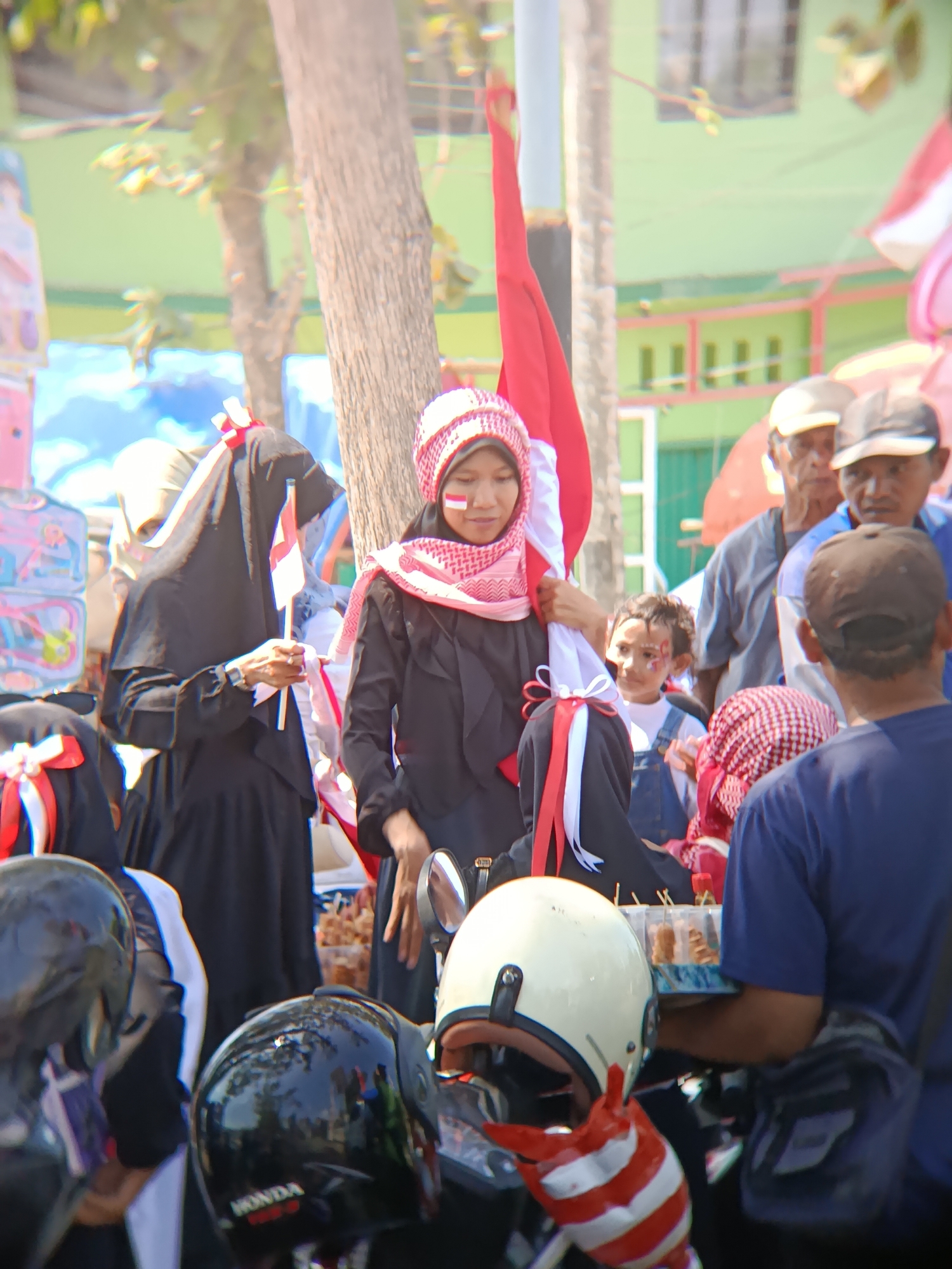 The image captures a daytime scene in Desa Kebon Manis, Kecamatan Cilacap Utara, Indonesia, featuring a gathering of people, many of whom are adorned with red and white face paint and attire, suggesting a patriotic or celebratory event. In the foreground, several motorcycle helmets are visible, including a black helmet with the "HONDA" logo. A young person, possibly a child, is seen with red and white face paint and denim overalls. Several adults are present, including a woman wearing a black hijab and holding small Indonesian flags, and another woman in a pink and white patterned hijab. The background shows buildings with green walls and a tree. The overall atmosphere appears to be lively and communal.