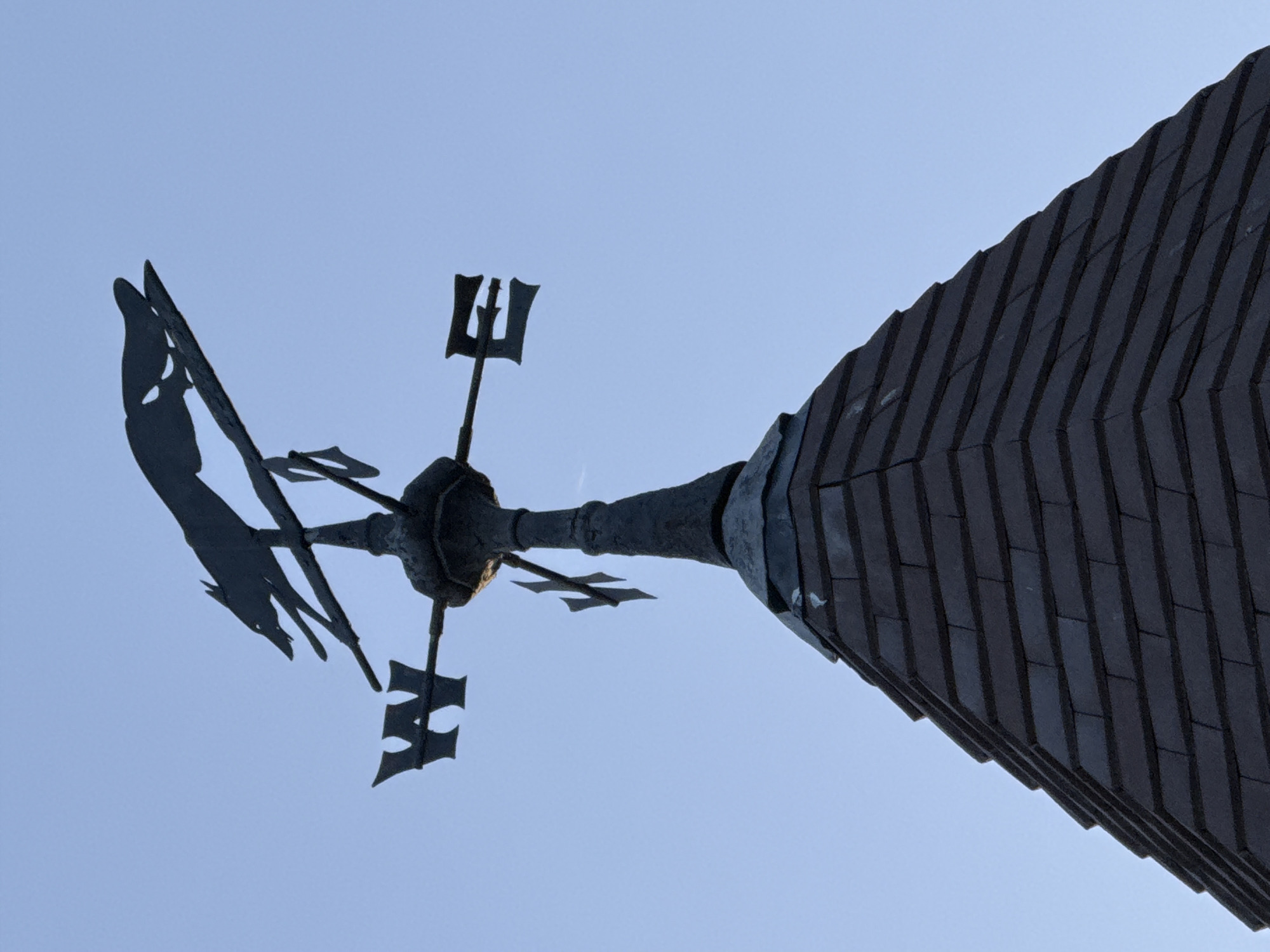 The image shows a low-angle view of the top of a building, most likely a church spire, with a weather vane against a light blue sky. The spire is covered in dark, rectangular tiles arranged in a regular pattern. The weather vane is metallic and features stylized cardinal directions (north, south, east, and west) as well as a rooster silhouette. The location is most likely in the city of London, United Kingdom, due to the architectural style commonly seen in historical buildings and churches. No people are visible.