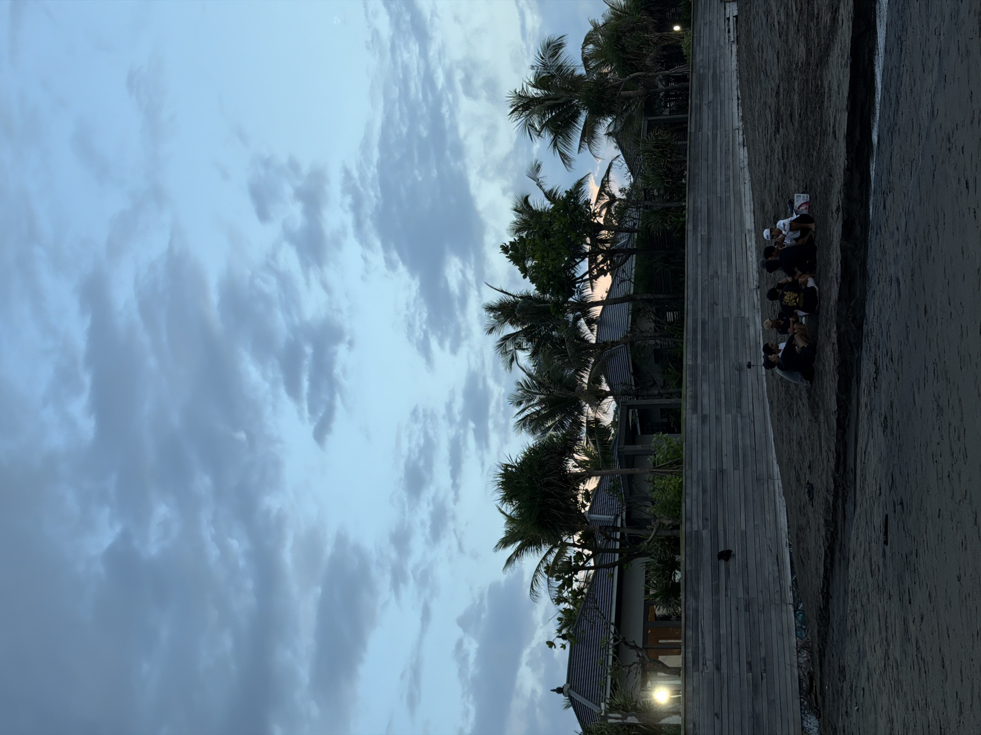 The image depicts a coastal scene in North Kuta, Indonesia. The sky is overcast with heavy clouds. A row of palm trees lines a wooden walkway. To the right of the walkway is a sandy beach where a group of people are sitting together. The buildings of a beach resort or villas can be seen behind the trees. A spotlight is illuminated near the buildings.