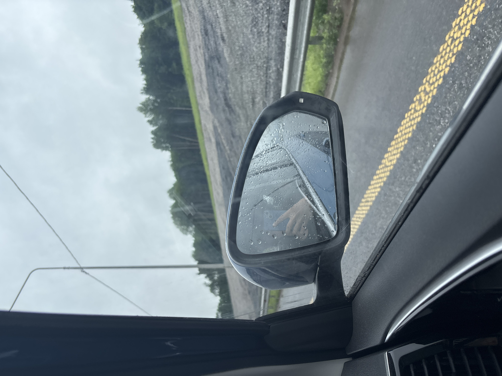 The image, taken from inside a vehicle, captures a view through the passenger-side window and side-view mirror, oriented on its side. When rotated to the correct upright position, it depicts a rainy or recently rainy day on a rural road in Savaleevo, Russia.
The central subject is the car's passenger-side wing mirror, heavily speckled with raindrops. Reflected in the mirror is a light-skinned hand, seemingly holding a smartphone or a similar small device. A small, square-shaped sensor or indicator light is visible on the top outer edge of the mirror housing.
Beyond the car, a wet asphalt road is visible, featuring a double solid yellow line composed of individual raised dots or bumps. Parallel to the road, a metal guardrail stretches into the distance. Past the guardrail and road, a wide, damp field or agricultural area extends towards a dense line of dark green trees that form the horizon. The sky is uniformly overcast and grey, indicating a cloudy or rainy day. Thin wires, likely utility lines, are faintly visible against the sky in the upper left background (before rotation).
The scene suggests the car is traveling or pulled over on a highway or rural road during inclement weather. The overall mood is quiet and muted due to the cloudy conditions and wet environment. No legible text is visible in the image.
