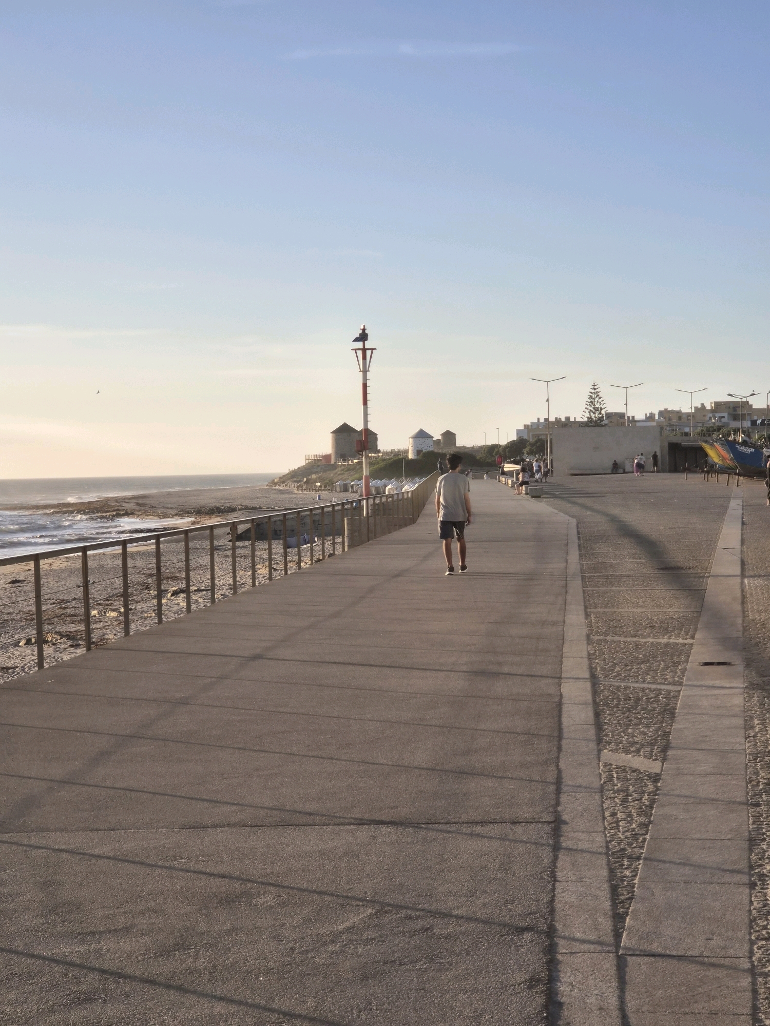 A young man walks away from the camera down a paved promenade that borders a beach. The promenade has a metal railing on the left separating it from the beach. The beach has rocks and some seaweed. The ocean is visible to the left, with waves breaking on the shore. In the background, there are buildings with windmills, streetlights, and boats. The sky is clear and blue with a soft gradient towards the horizon. The scene is likely the waterfront in Esposende, Portugal.