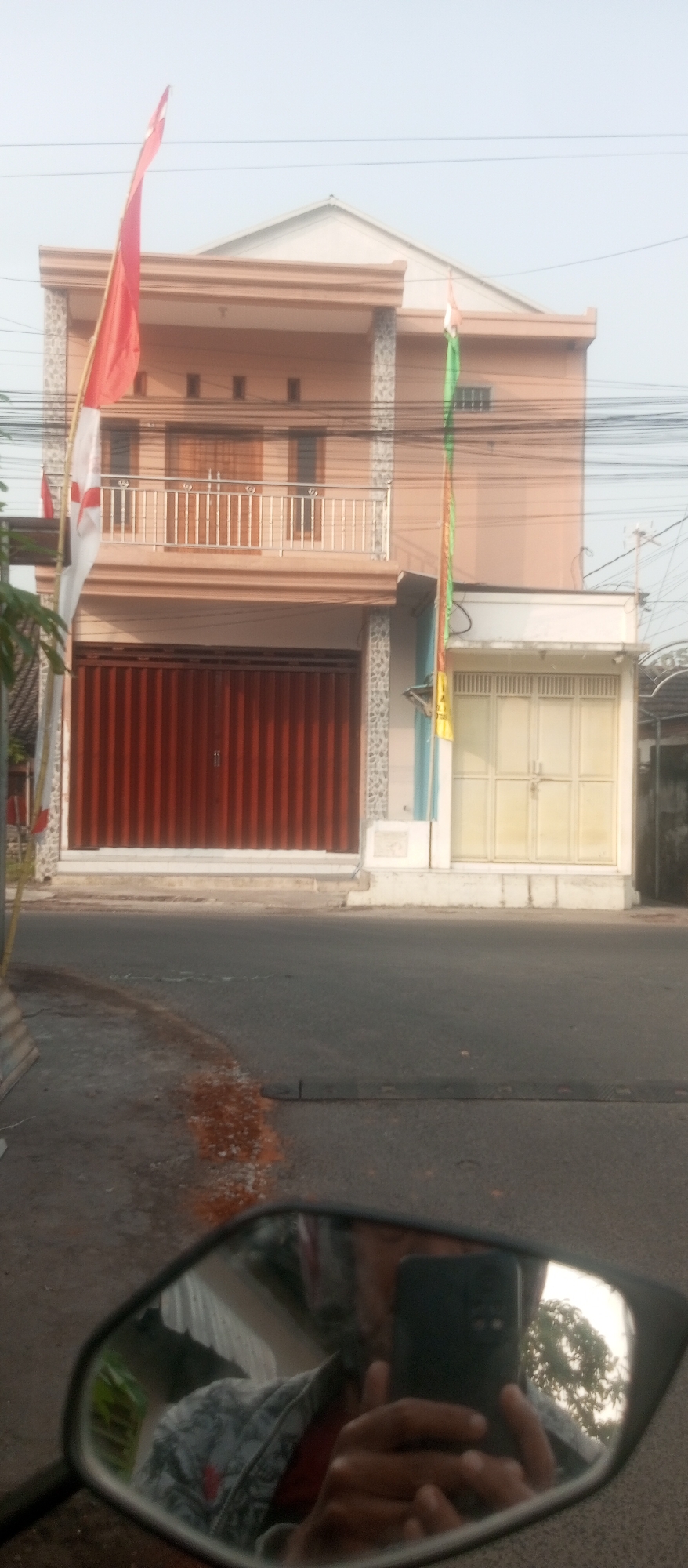The image captures a street scene in the city of Kediri, Indonesia. In the foreground, a motorcycle's side mirror reflects a person holding a dark-colored smartphone, and their upper body is visible wearing a grey patterned jacket over a red shirt. The background features a two-story building with peach-colored walls. The ground floor has a large, closed, reddish-brown metal roller shutter door. Above this shutter, there's a balcony with a silver railing and two windows. A red and white Indonesian flag flies from the left side of the building, and a green, white, and orange flag is attached to the right side. To the right of the main building, a smaller section with cream-colored double doors is visible. The street in front of the building has a faded orange marking and some scattered white debris. Overhead, power lines are strung across the frame. The sky is overcast and light-colored, suggesting a daytime scene.