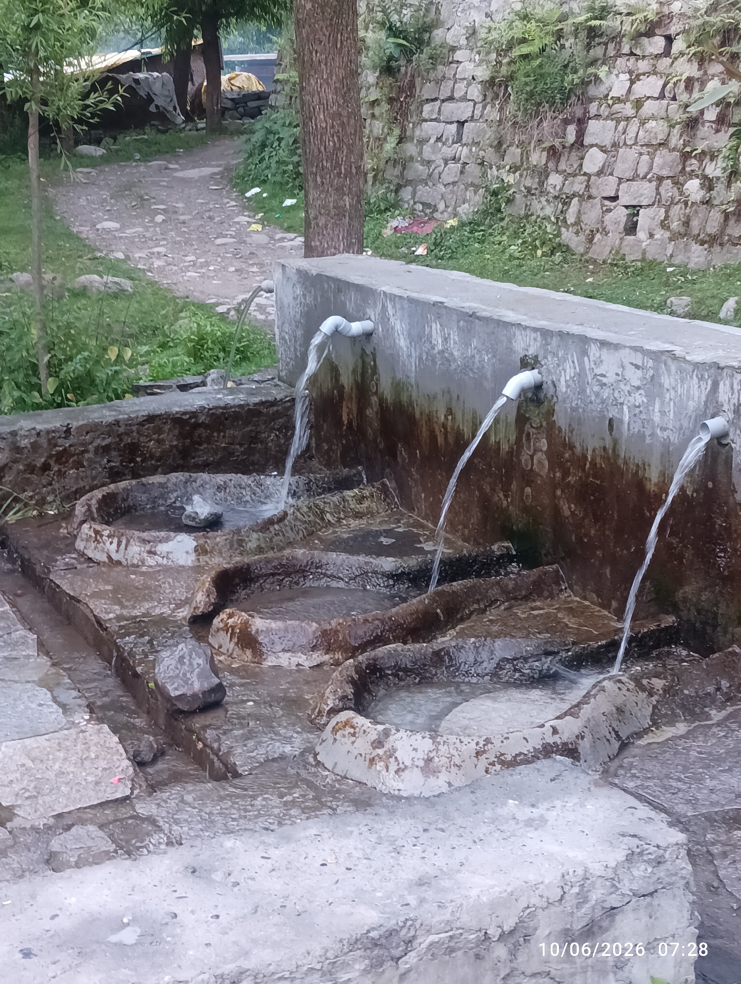 This image depicts an outdoor water fountain in Goshal, India. The fountain is constructed from concrete and features three spouts from which water is flowing into carved basins. The basins appear to be shaped for handwashing or filling containers. The surrounding environment includes a stone wall behind the fountain, overgrown vegetation, and a dirt path leading away into the distance. The ground in front of the fountain is paved with stone slabs, some of which are wet from the water. The overall scene suggests a functional, public water source in a natural or semi-rural setting.