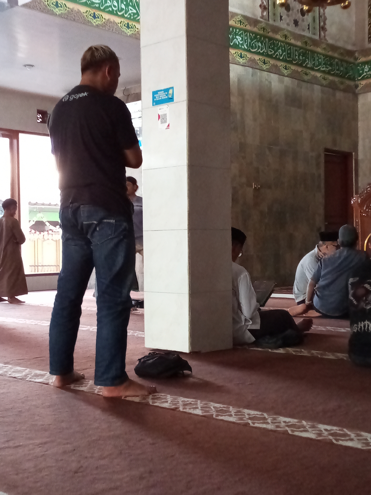 The image depicts the interior of a mosque in Sariwangi, Indonesia. In the foreground, a man wearing a black t-shirt with a "gojek" logo and blue jeans stands facing away from the camera. His back is slightly turned, and he is looking towards the right of the frame. His feet are bare, resting on a red carpeted floor marked with white decorative lines. To his right, a large, square pillar is visible, with a white notice attached to it.
Further into the mosque, several other individuals are present, some sitting on the carpet. One person, wearing a light-colored robe, is partially visible on the left. Towards the back of the frame, a group of men are seated on the floor. One man in the center is wearing a white shirt and a black cap, appearing to be looking down. To his right, another person in a blue t-shirt and a grey cap is visible, also seated. A black bag lies on the floor near the pillar. The walls of the mosque are adorned with decorative Islamic calligraphy and patterns. The overall lighting suggests an indoor environment, likely during daytime.