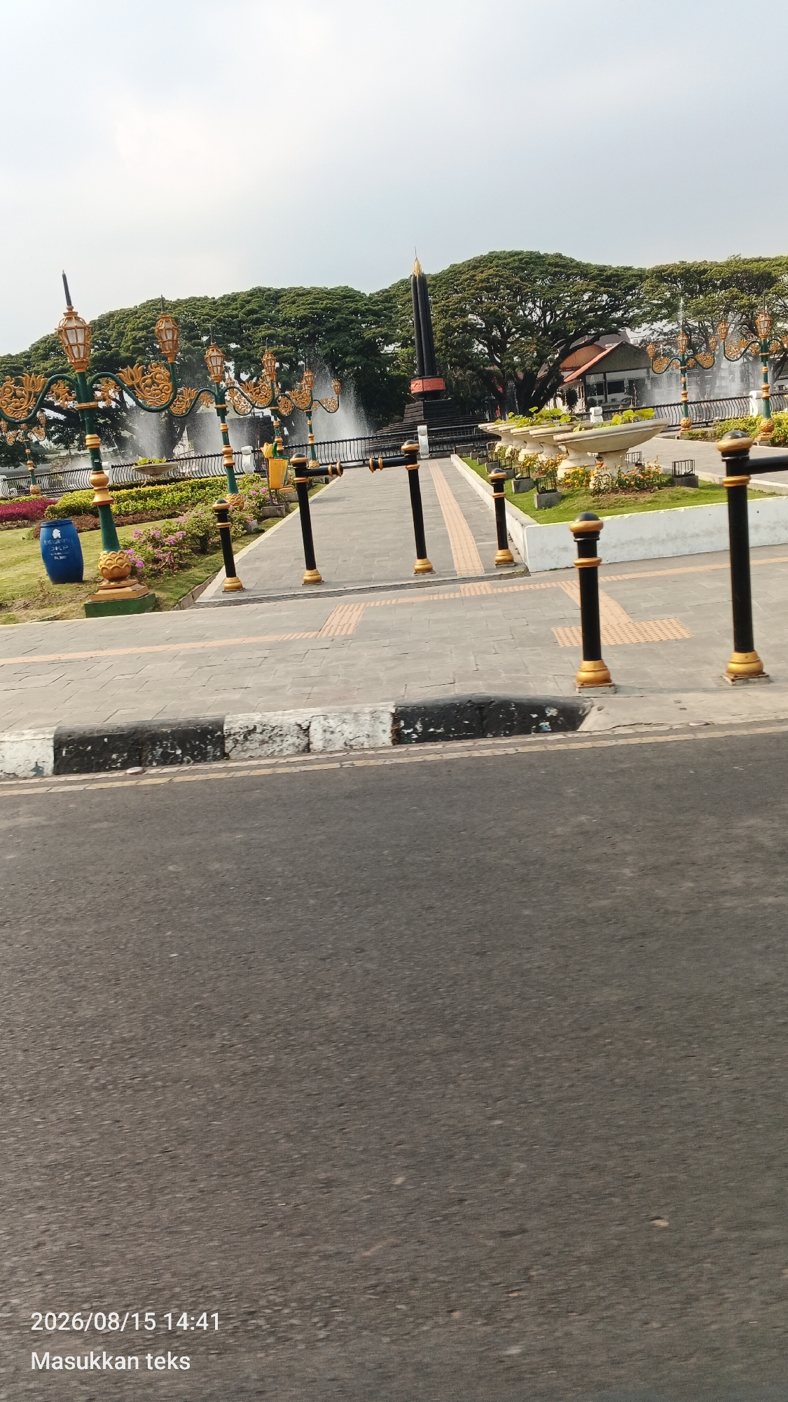 The image depicts an outdoor scene in the city of Kiduldalem, Indonesia, likely a park or public square. In the foreground, a paved road with a white and black painted curb is visible. Beyond the curb is a paved walkway lined with decorative black and gold bollards. The walkway leads towards a park area with lush green trees, fountains spraying water, and ornamental street lamps with gold accents. A tall, dark obelisk-like monument stands prominently in the background, surrounded by greenery and a building. There are no people or animals visible in the scene. The sky is overcast. A blue trash bin is also present in the park area. The image also has a timestamp overlaid in the bottom left corner: "2026/08/15 14:41" and the text "Masukkan teks".