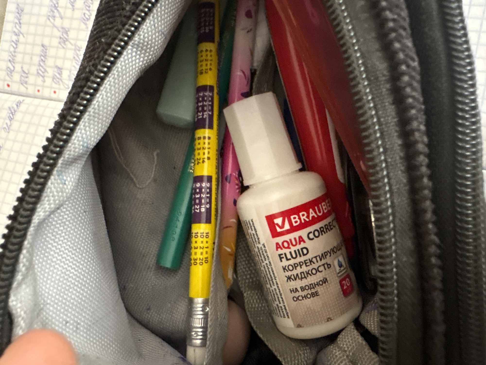 The image shows the inside of a dark grey pencil case. Several writing implements are visible, including a yellow pencil with multiplication tables printed on it, a green pencil, a pink pencil, and red pens. A small white bottle of correction fluid with a red label that says "AQUA CORRECTION FLUID" is also present. The correction fluid bottle has text in Russian, indicating it is "corrective fluid on a water base." The pencil case appears to be made of fabric with a zippered closure. The background shows a piece of paper with handwritten text and a grid pattern, suggesting it is a notebook. The scene is lit from above, casting shadows within the pencil case. There is no discernible location in Georgiyevsk, Russia.