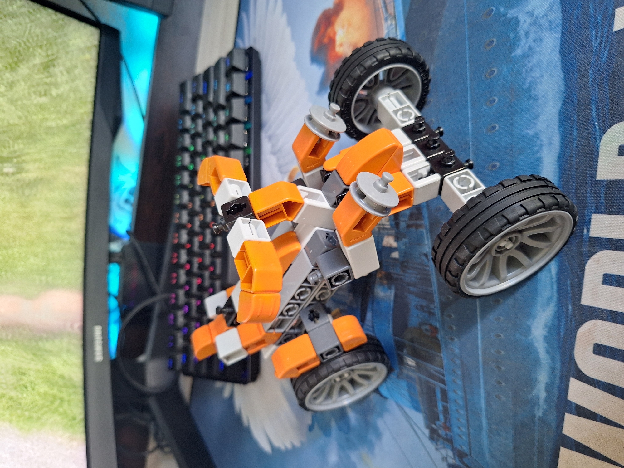 An orange and white LEGO ATV is positioned on a desk with a blue and white ship-themed mousepad. In the background, a black keyboard with illuminated keys and a black monitor displaying a green image are visible. The scene appears to be indoors. There is no indication of the city of Varna, Bulgaria in the image.