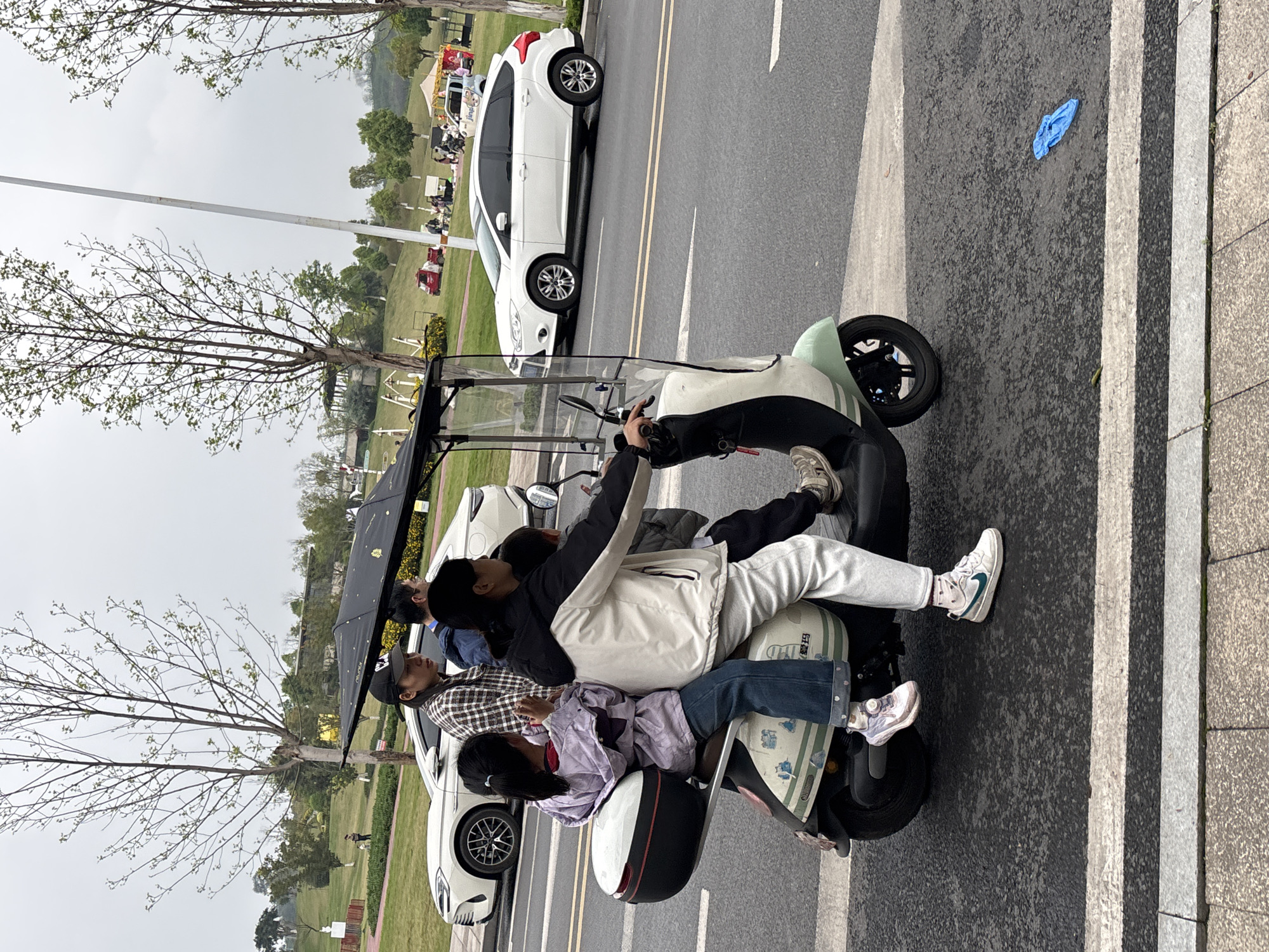 An electric scooter, light green and white with a black sunshade canopy and a rear storage box, is captured in motion on a paved road in Jiangjin, China. Four individuals are riding the scooter: an adult male driving, wearing a black and white jacket and white sneakers; a young boy positioned in front of the driver, obscured; an adult female seated behind the driver, wearing a plaid shirt and black baseball cap; and a young girl seated at the rear, wearing a purple jacket and blue jeans, holding a small red object. The road features double yellow lines and white crosswalk markings. To the right of the scooter, a white BYD Seal U / Song Plus EV and a white Ford Focus hatchback are visible on the road. The background reveals a public park environment with grassy hills, numerous trees with budding leaves, and an overcast sky. In the mid-ground of the park, several individuals are gathered around small tents or stalls, and a red pickup truck is parked. A white utility pole stands centrally in the background. In the distant left, a person walks a dog near a white statue and red fencing. A blue disposable glove lies on the asphalt in the lower right foreground, adjacent to a concrete sidewalk.