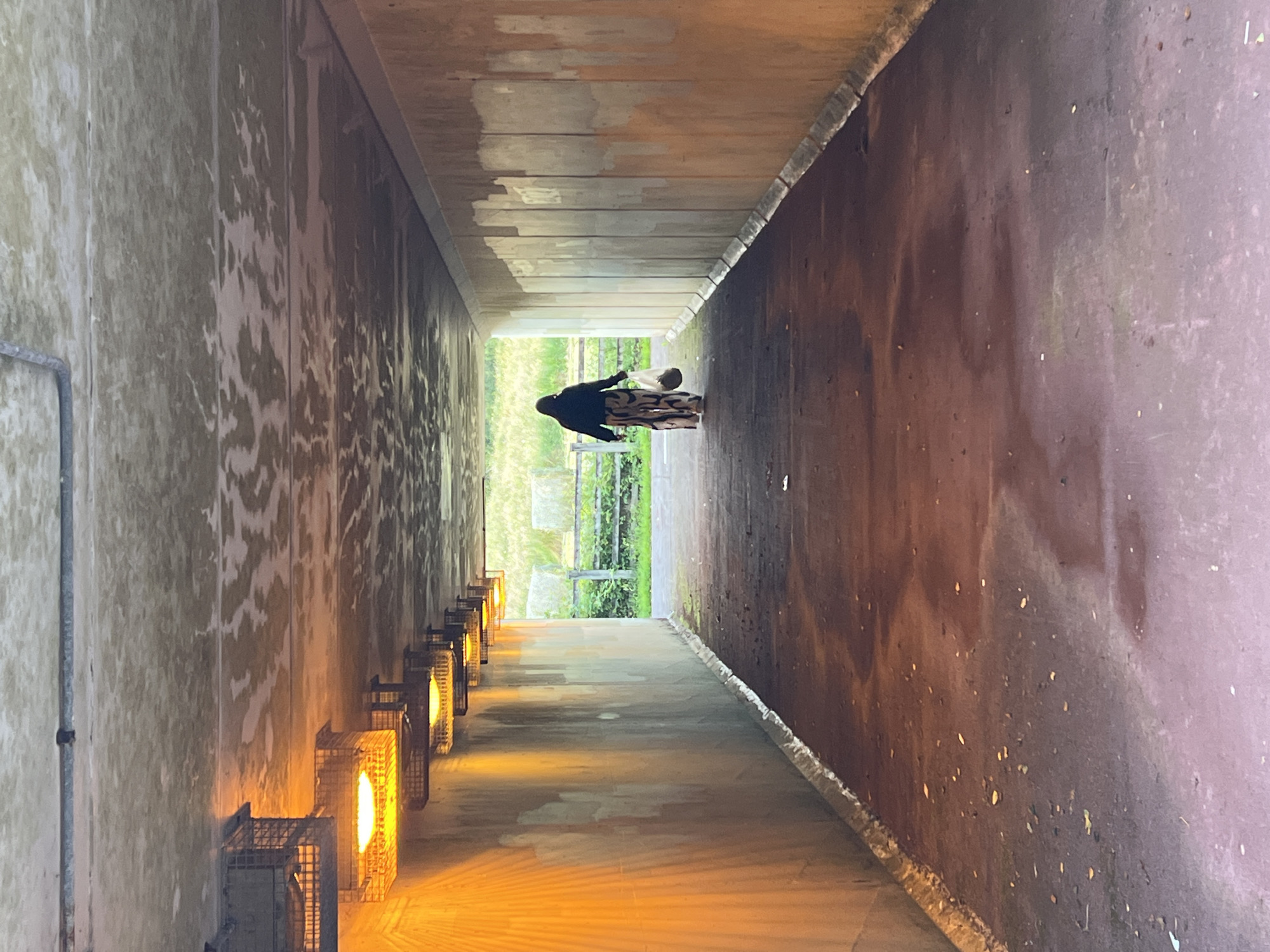 The image shows a person walking through a tunnel in Mountcharles, Ireland. The tunnel has concrete walls and a ceiling with lights mounted along the sides. The person is wearing a dark hooded jacket and patterned trousers and is carrying two white plastic bags. The tunnel floor is a dark, reddish-brown color and is littered with some debris. The tunnel opens into an area with a grassy field and a fence in the background. The lighting in the tunnel is dim and warm, emanating from the overhead fixtures.