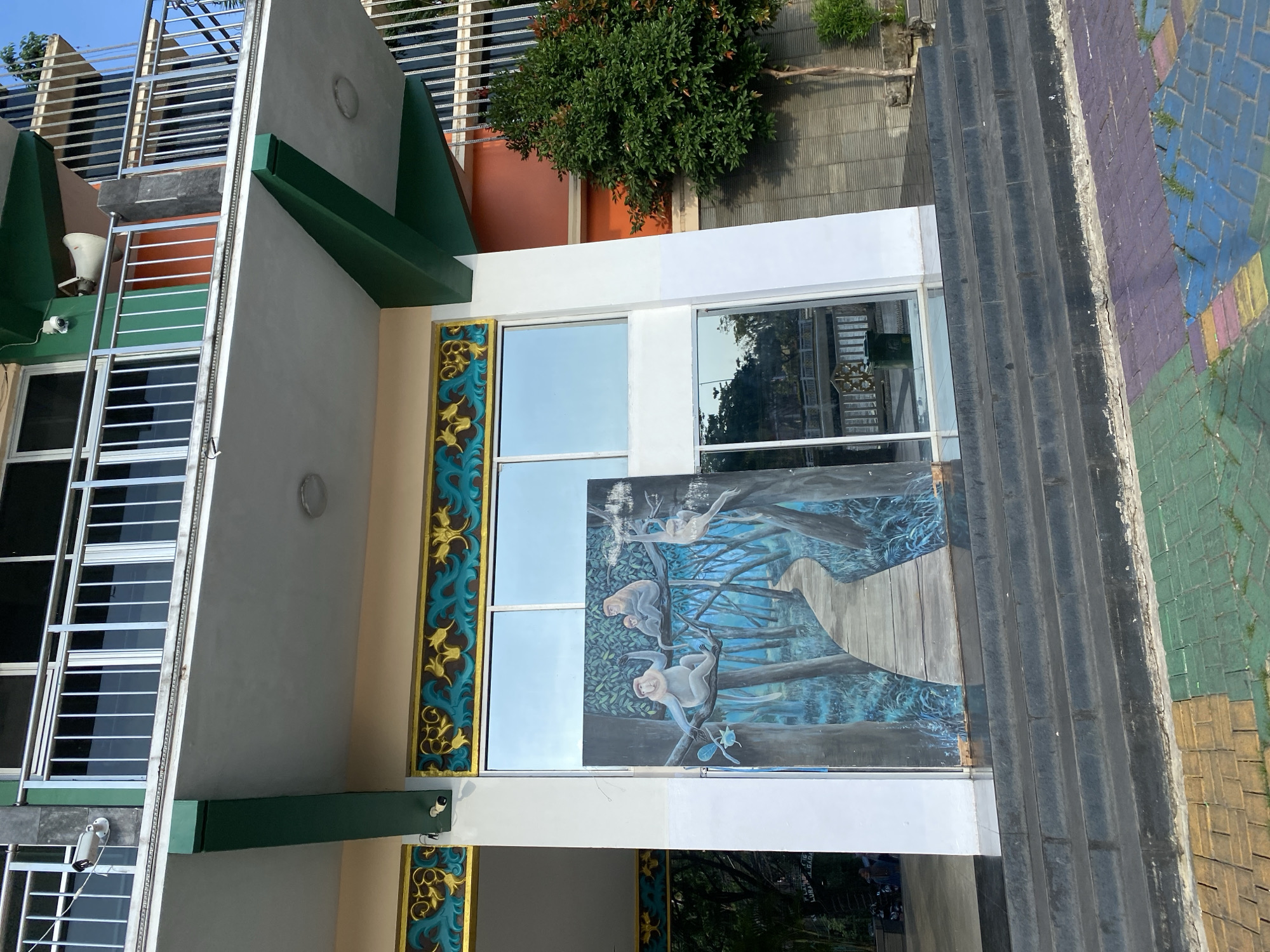The image shows the exterior of a building with a mural of monkeys in a forest setting. The building has multiple stories, with balconies and windows. There are decorative elements above the windows, including gold and teal patterns. A security camera is visible on the side of the building. In front of the building, there are stairs leading up to the entrance, and the ground is paved with colorful tiles. A small tree is planted to the right of the building. The location is identified as the Monkey Forest Sanctuary in Banjarmasin, Indonesia. There are no people or animals directly visible in the main part of the image, though motorcycles are visible in the background to the left. The lighting suggests it is daytime, possibly late afternoon due to the shadows.