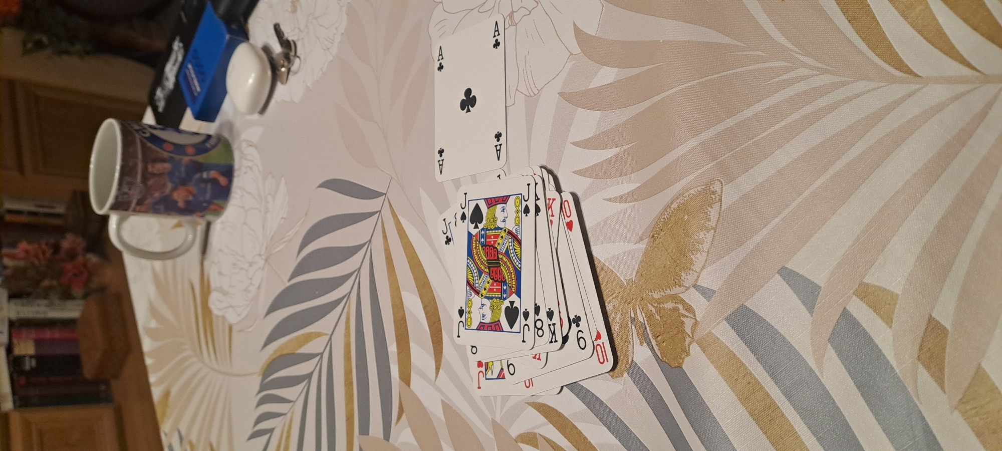 A tilted close-up shot displays a collection of playing cards and other items scattered on a table with a patterned tablecloth. The playing cards include the Ace of Clubs, a Jack of Spades, and others in an incomplete sequence ending with the 6 of Hearts. A blue container, a white oval object, and keys are positioned above the cards, closer to the top-left corner. A ceramic mug with a photographic image sits nearby, also near the top-left. The tablecloth features a pattern of stylized leaves and a butterfly. The setting is indoors. There is no visual evidence to suggest the location is Burgas, Bulgaria.