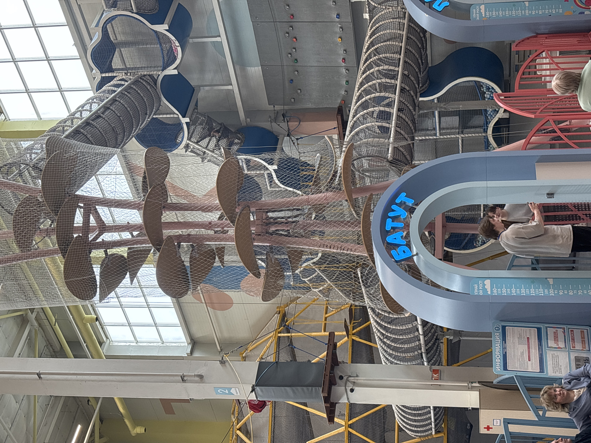 This image depicts a vibrant, multi-level indoor entertainment complex, likely a trampoline park or adventure playground, located in Perm, Russia. The scene is characterized by high ceilings, ample natural light from large, grid-patterned windows, and a multitude of interconnected play structures.
In the foreground, two prominent light-blue archways with dark blue trim stand, one displaying the Cyrillic word "БАТУТ" (Trampoline) in glowing blue letters. Attached to the right arch is a white sign with a blue border, serving as a height chart ranging from 50 to 160 cm, labeled "УЗНАЙ СВОЙ РОСТ!" (Find your height!). To the left of the archways, an information stand is visible, also blue-framed and featuring the word "ИНФОРМАЦИЯ" (Information). Red metal gates are situated behind the archways.
Several individuals are present. In the lower-left foreground, a woman with blonde hair, wearing a light-colored top, is seated, looking towards the right. Another person is partially visible next to her. Near the right archway, a young person in a light-colored long-sleeved shirt stands, looking towards the right, seemingly interacting with another person whose back is partially seen.
The complex features a dominant central climbing tower with a reddish-pink central column, from which numerous brown, mushroom-shaped platforms extend, interconnected by white safety netting. Various blue and grey netted tunnels and slides crisscross the expansive space at different elevations. One blue, cloud-shaped section of a slide contains numerous white and blue plastic balls, indicating a ball pit. To the left, a section of a yellow-framed rope course or high-level obstacle course, also netted, is visible, supported by white structural pillars. A red bag hangs from a rope on this course. On the far right wall, a gray climbing wall is equipped with multiple colored handholds (red, blue, green, orange). Figures of individuals are discernible within the upper levels of the netted structures and tunnels, indicating active participation. The overall environment appears well-lit, clean, and designed for recreational activities.