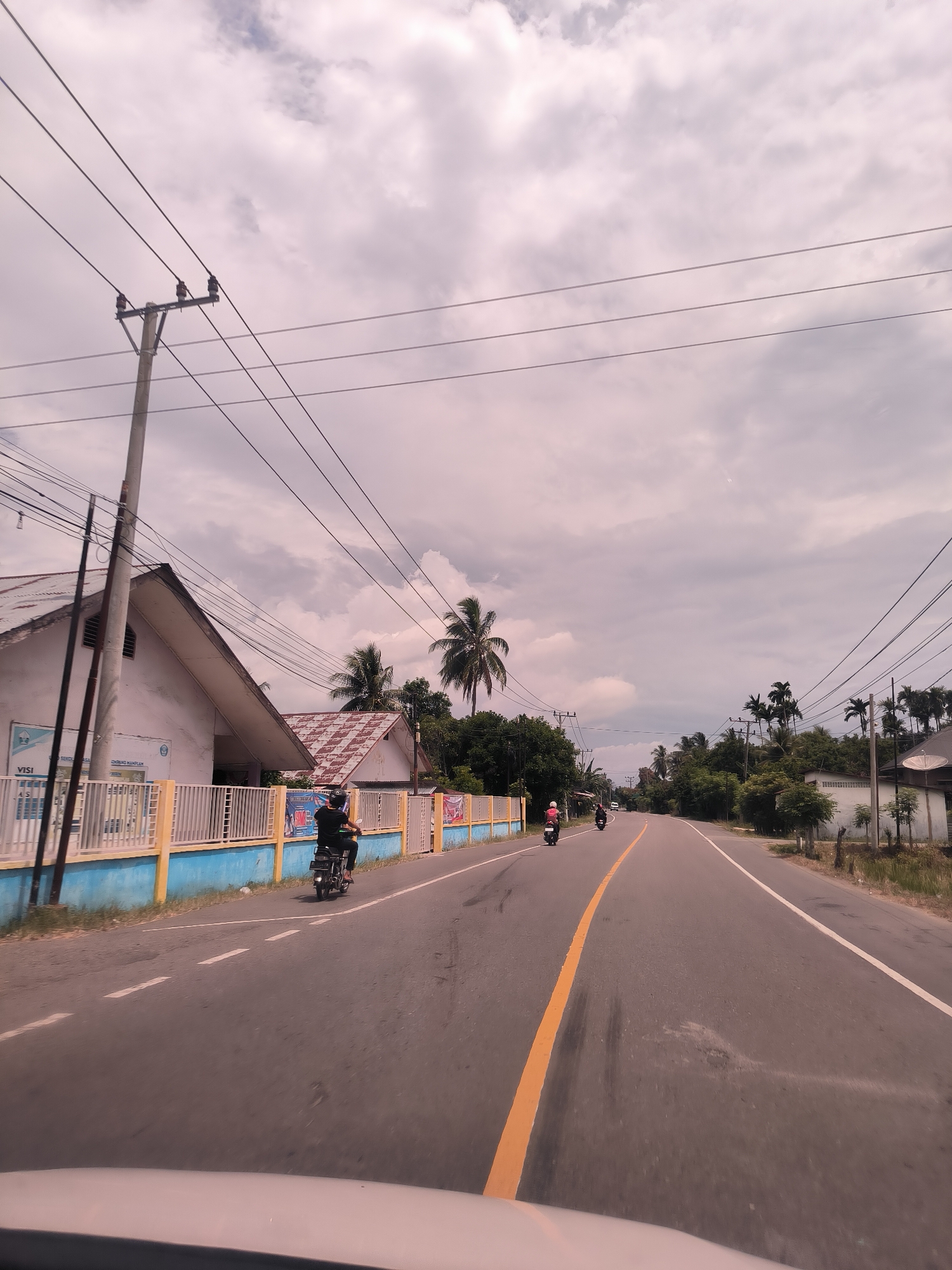 A wide-angle, eye-level shot captures a paved road stretching into the distance in a semi-rural setting in Ierhobglumpang, Indonesia, under a cloudy, overcast sky. The foreground reveals the top of a vehicle's dashboard, indicating the photo was taken from inside a moving car.
The asphalt road features a clear yellow solid line marking the center and white dashed lines on the shoulders. Three motorcycles are visible on the road. On the left shoulder, a rider in a black shirt and helmet is either stopped or moving slowly. Further ahead, two more motorcycles are seen, with one rider wearing a pink helmet.
On the left side of the road, a prominent concrete utility pole with multiple power lines stands against the sky. Behind it, a white house with a steep, light-colored tiled roof is partially obscured by a bright blue and yellow fence. Text visible on the fence includes "VISI" and "PENDING HARAPAN," along with part of a banner that reads "SMAN." Another house with a reddish-brown corrugated roof is nestled behind the first building, surrounded by lush tropical foliage.
Both sides of the road are lined with abundant greenery, dominated by numerous tall palm trees. Further down the road on the right, more residential structures and a white building with a satellite dish are visible. The time of day appears to be daytime, with diffused light due to the heavy cloud cover. The scene depicts typical daily activity along a local road in this Indonesian context.