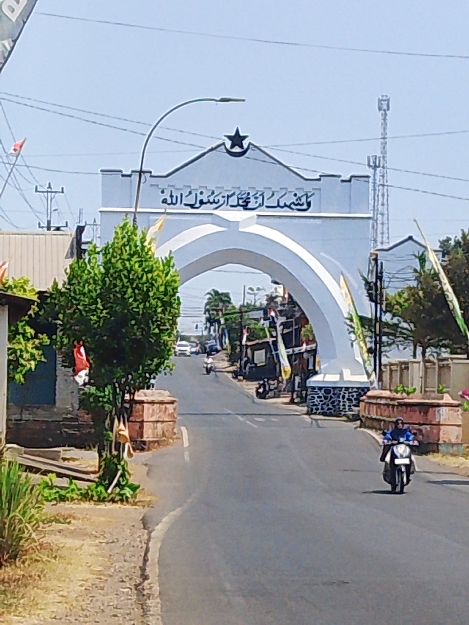 The image shows a street scene in Mantingan, Indonesia, under a clear sky. A large, white archway, adorned with Arabic script and a star, dominates the center of the frame. The archway is supported by stone pillars. To the left of the archway, a small tree with dense green foliage stands on a brick pedestal. A red and white Indonesian flag is visible hanging from a pole near the tree. The street leads away from the viewer, under the arch. Several cars and a motorcycle are visible on the road, with people riding the motorcycle. Buildings with corrugated metal roofs are present on either side of the street. The environment appears to be a suburban or semi-rural area with moderate vegetation.