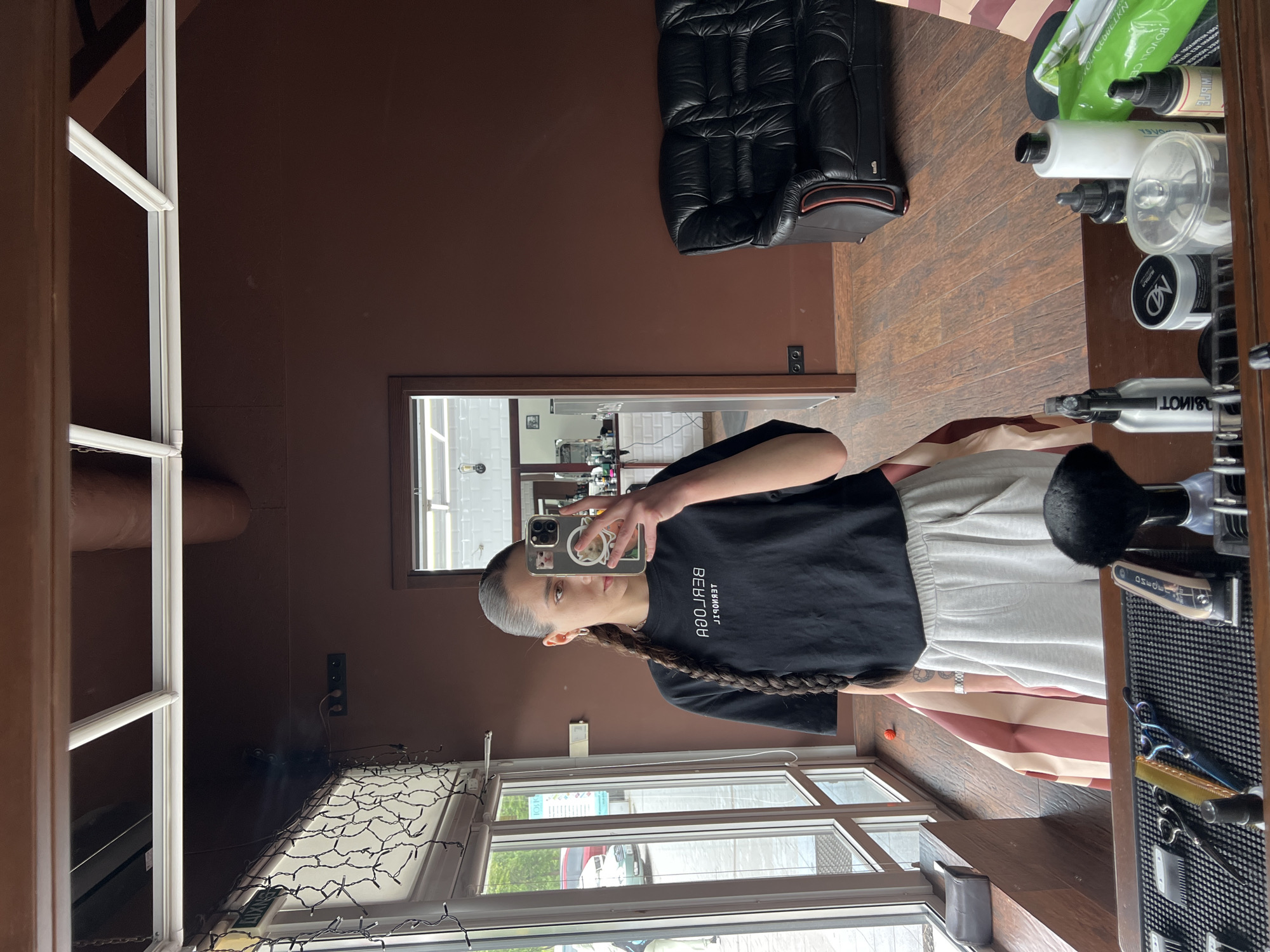 A woman with a long braid is taking a selfie in what appears to be a hair salon. The salon has dark brown walls and wood flooring. There is a black quilted chair in the background, along with shelves stocked with various hair care products. A window with a view of the outside is visible, suggesting a possible street or urban setting. The woman is wearing a black t-shirt and light-colored pants. There's a barber's station with clippers, scissors, and brushes nearby. Based on the context, the location is possibly in Ternopil, Ukraine.
