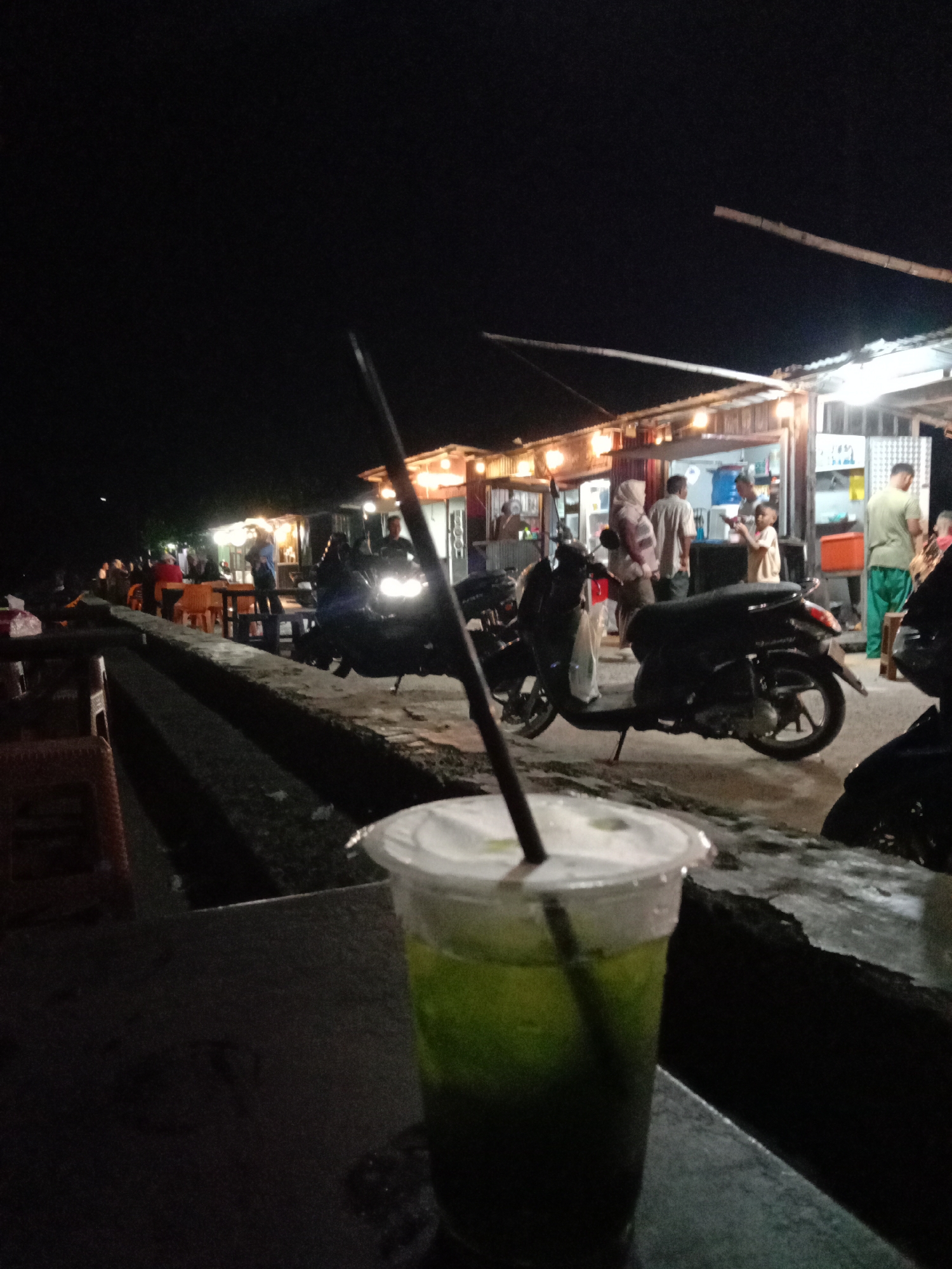 The image captures a lively night scene in Lhokseumawe, Indonesia, characterized by artificial lighting against a dark sky. In the immediate foreground, a clear plastic cup filled with a vibrant green beverage, possibly a matcha drink or similar, with a straw and white lid, sits on a dark, textured surface, likely a table or ledge.
The midground and background reveal an outdoor market or street food area bustling with activity. A line of small, illuminated stalls or shops extends into the distance on the right, casting bright light on the surrounding area. These structures appear to be constructed from simple materials like corrugated metal or wood. Several people are visible around these stalls, some standing, some seated at tables with orange chairs, suggesting they are customers purchasing food or drinks, or vendors attending to their businesses.
Multiple motorcycles or scooters are parked along the path, indicating a common mode of transport in the area. A low concrete barrier or wall runs from the foreground along the left side of the scene, separating the immediate viewing area from the activity beyond. The strong contrast between the brightly lit stalls and the dark night sky creates a dynamic atmosphere. No discernible text is clearly visible on any signs or objects. The overall impression is one of casual evening commerce and social gathering in a tropical setting.