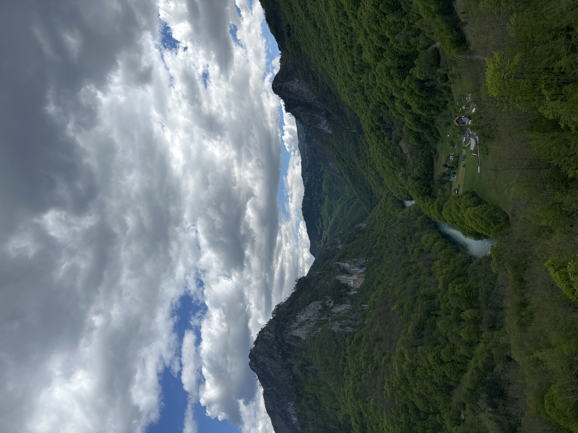 The image depicts the Tara River Canyon in Đurđevića Tara, Montenegro, with the river flowing through a deep gorge flanked by steep mountains covered in lush, green forest. On the left, a craggy, gray mountain rises, partially obscured by dramatic, billowing clouds. The sky is a mix of white clouds and patches of blue. There is a settlement with several small buildings and open areas near the river on the right side of the canyon.