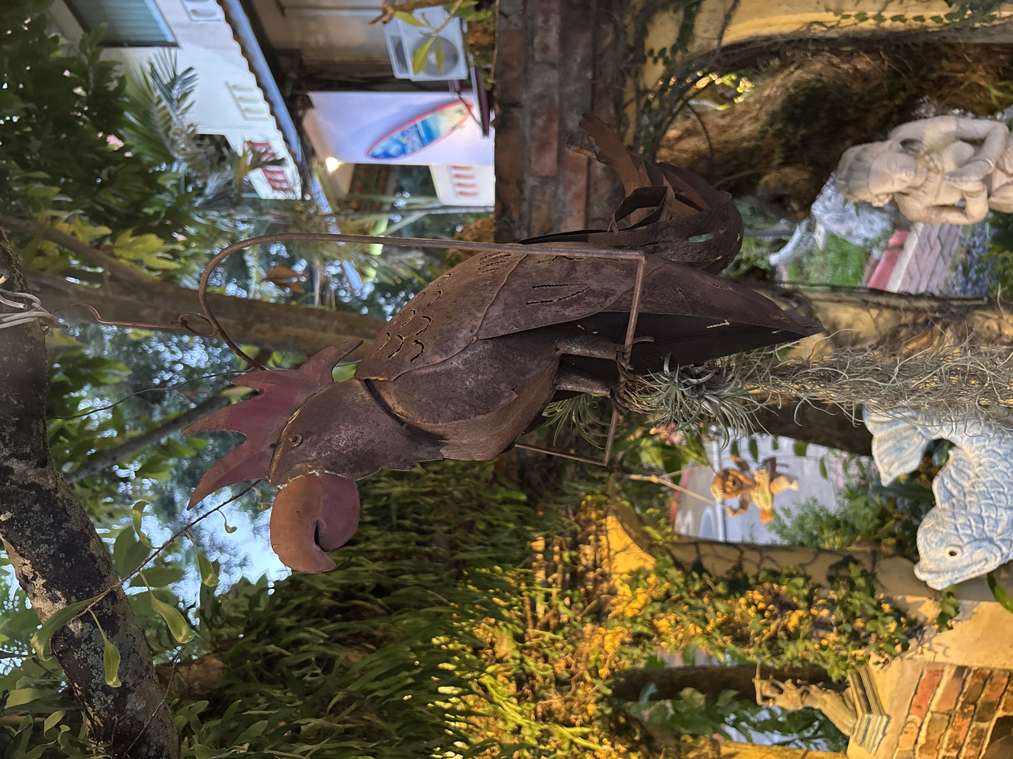 This image depicts an outdoor decorative scene, likely in Chalong, Thailand. In the foreground, a rusty metal sculpture of a parrot hangs from a tree branch. The parrot faces left and is intricately detailed, with carved feathers and a prominent beak. Below the parrot, a light blue ceramic fish sculpture is partially visible. To the right, a light-colored statue of a kneeling woman with her hands pressed together is situated. The background reveals a white building with red-trimmed windows and a portion of a white banner with a surfboard graphic. Lush green foliage and trees surround the area, illuminated by soft, warm lighting, suggesting it may be evening or early morning. The overall setting appears to be an outdoor courtyard or garden area, possibly part of a restaurant or resort.