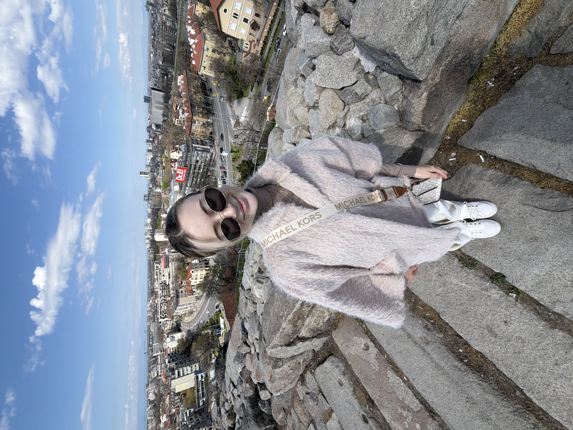 A young woman stands on the Nebet Tepe hill in Plovdiv, Bulgaria, against a backdrop of the city. She wears a light pink, furry jacket with a Michael Kors crossbody bag and white sneakers. The city skyline is visible in the background, with buildings and streets stretching into the distance under a blue sky with scattered clouds. The ground is made of stone.