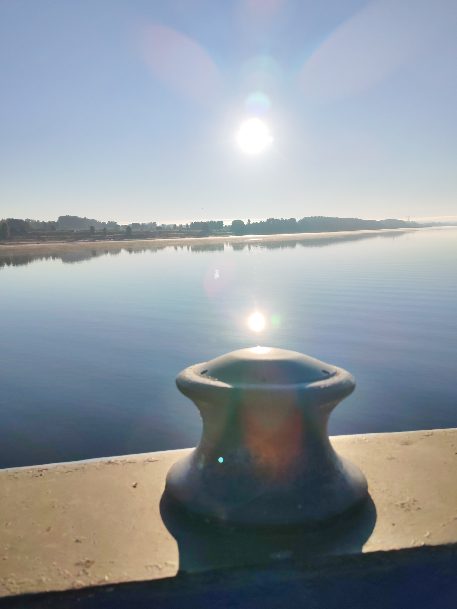 This image captures a serene morning scene by a body of water, likely a river or lake, in Bol'shie Mkhi, Russia. The sun, low in the sky, is intensely bright and causes lens flare, creating a circular halo effect and a diffused glow across the upper portion of the image. The water's surface is calm and reflects the pale blue sky and the distant treeline. A thin layer of mist or fog hovers just above the water near the shore, softening the landscape.
In the foreground, a dark, weathered bollard stands on a concrete ledge. The bollard has a distinctive shape with flared ends, suggesting it's used for mooring boats. The concrete ledge is rough and shows signs of age and exposure to the elements. The overall atmosphere is tranquil and peaceful, emphasizing the quiet beauty of the natural surroundings as dawn breaks. No people are visible in the frame.