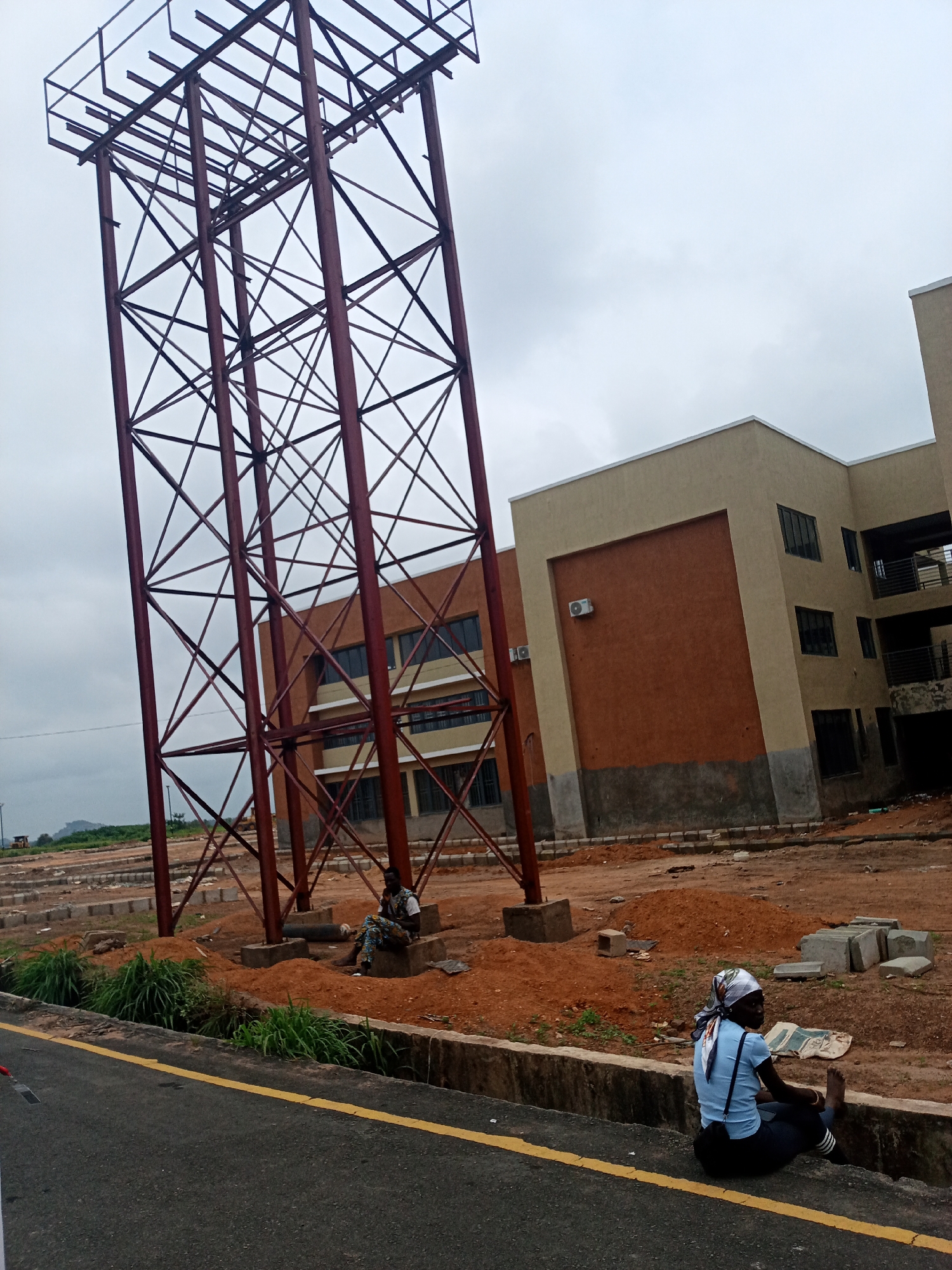 This image depicts an outdoor construction scene in Iseyin, Nigeria. Dominating the foreground is a tall, reddish-brown metal lattice structure, possibly part of a billboard or telecommunications tower, reaching towards a cloudy sky. To the right of this structure, a modern building with beige and brown facades and dark-framed windows is under construction. The ground is a mix of reddish-brown earth, gravel, and concrete blocks.
In the mid-ground, two individuals are present. One person, a male wearing a white shirt and patterned trousers, is seated on a concrete block at the base of the metal structure. He appears to be holding a phone to his ear. Another person, a female wearing a light blue shirt, dark trousers, and a patterned headscarf, is seated on the edge of a concrete curb along the side of an asphalt road in the foreground. She is looking away from the camera.
The overall environment suggests an active construction site with a new building and a large metal frame. The weather appears overcast. The scene is set against a backdrop of undeveloped land with some vegetation and distant hills.