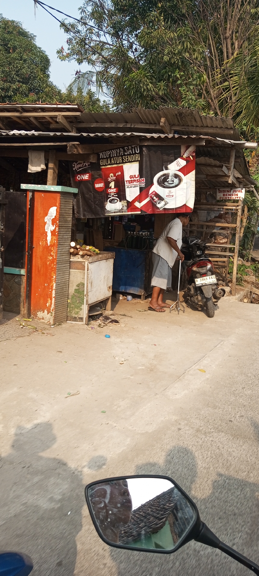 In Setu, Indonesia, a street vendor operates a small roadside stall. The stall features a corrugated metal roof supported by wooden beams and posts. Large banners advertise "Kopinya Satu Gula Atur Sendiri" (One Coffee Sugar Adjust Yourself) and "Gula Terpisah Jelas Lebih Enak" (Separate Sugar is Clearly Tastier), along with images of coffee. To the right of the stall, a sign reads "DISINI JUAL BENSIN PERTALITE" (Pertlite Gasoline Sold Here).
In front of the stall, a middle-aged person wearing a light-colored shirt and shorts stands with a walking cane, facing the stall. A red motorcycle is parked next to the person. In the foreground, various items are visible: a large orange structure on the left, a white counter with some produce and bottled liquids, and a blue cabinet. The ground is paved with concrete and appears dusty, with some debris scattered around. A reflection in a motorcycle rearview mirror shows the face of the person. The surrounding environment is characterized by trees and a bright sky, indicating an outdoor, possibly rural or semi-urban setting.