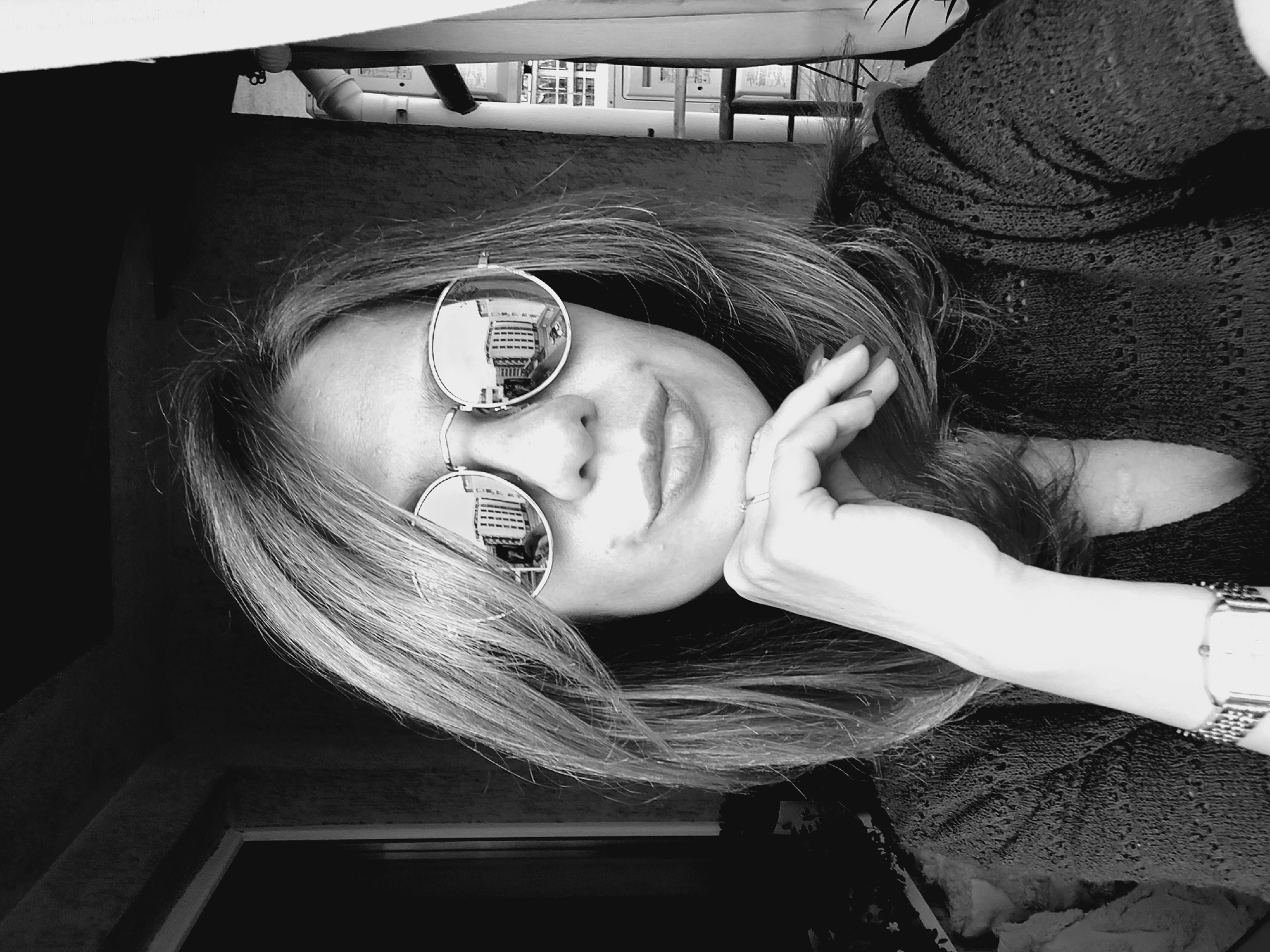 The black and white image is a close-up selfie of a person wearing round, reflective sunglasses. The person's face is tilted, and one hand is resting on their cheek. Their hair is long and appears to be light brown or blonde. The sunglasses reflect a building with multiple floors, possibly an apartment complex or office building. The setting appears to be outdoors, with some of the background suggesting a balcony or rooftop. The person is wearing a dark, textured sweater. A watch with a metallic band is visible on their wrist. The image seems to be taken during the daytime, as indicated by the lighting. The overall mood of the image is relaxed and perhaps a bit contemplative. No visible text is present in the image. The location context is Sofia, Bulgaria.