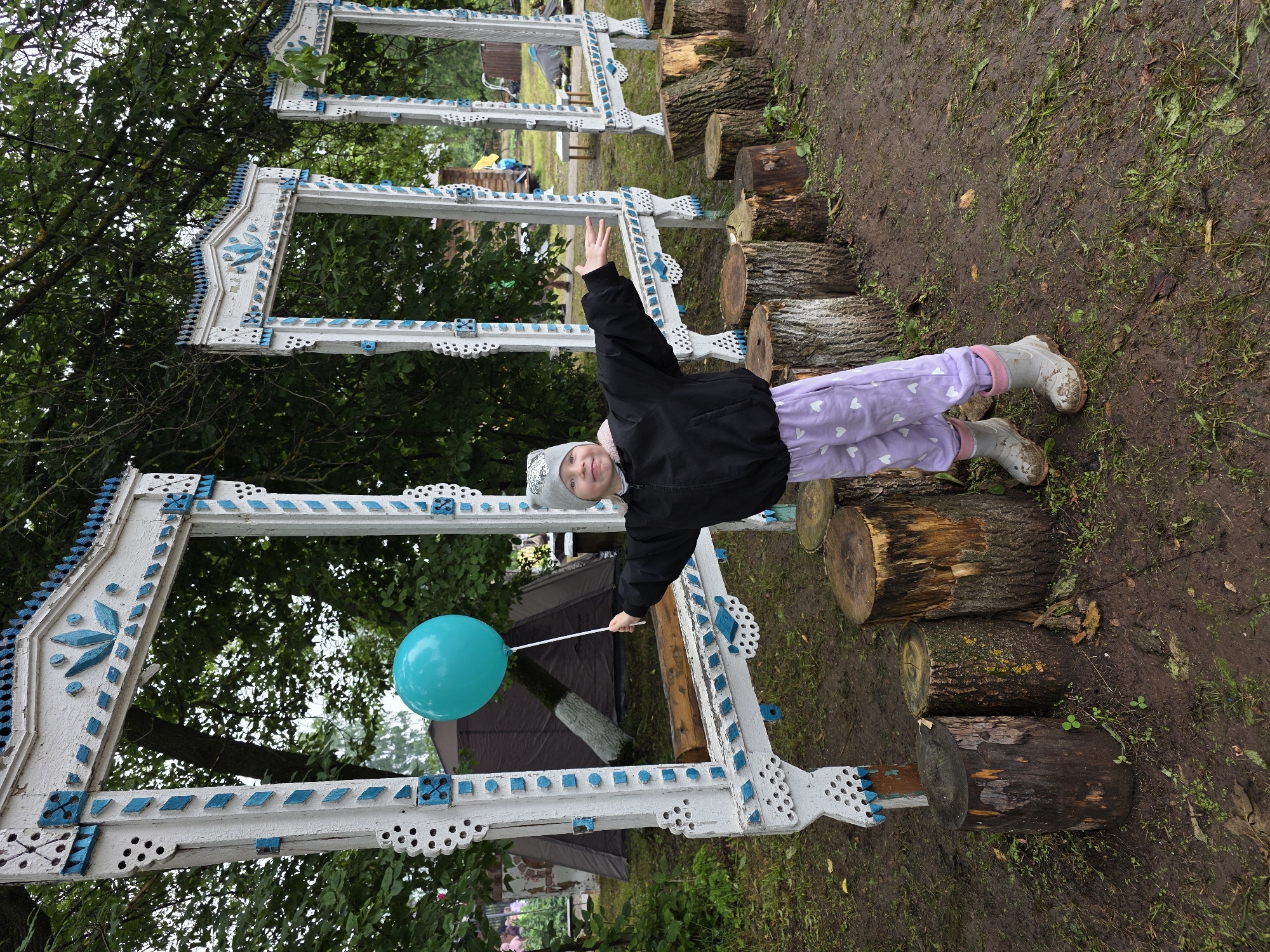 A young child stands outdoors, holding a teal balloon on a stick. The child is wearing a black jacket, light purple pants with white heart patterns, and grey boots. They are positioned in front of three ornate, white and blue decorative frames, which appear to be part of an outdoor installation. Behind the frames, a large, dark tent is visible, along with trees and foliage. The ground is muddy and covered with grass and fallen leaves, suggesting a natural or park-like setting. The overall scene appears to be in a rural location, possibly the village of Staroye Bygi, Russia, given the context.