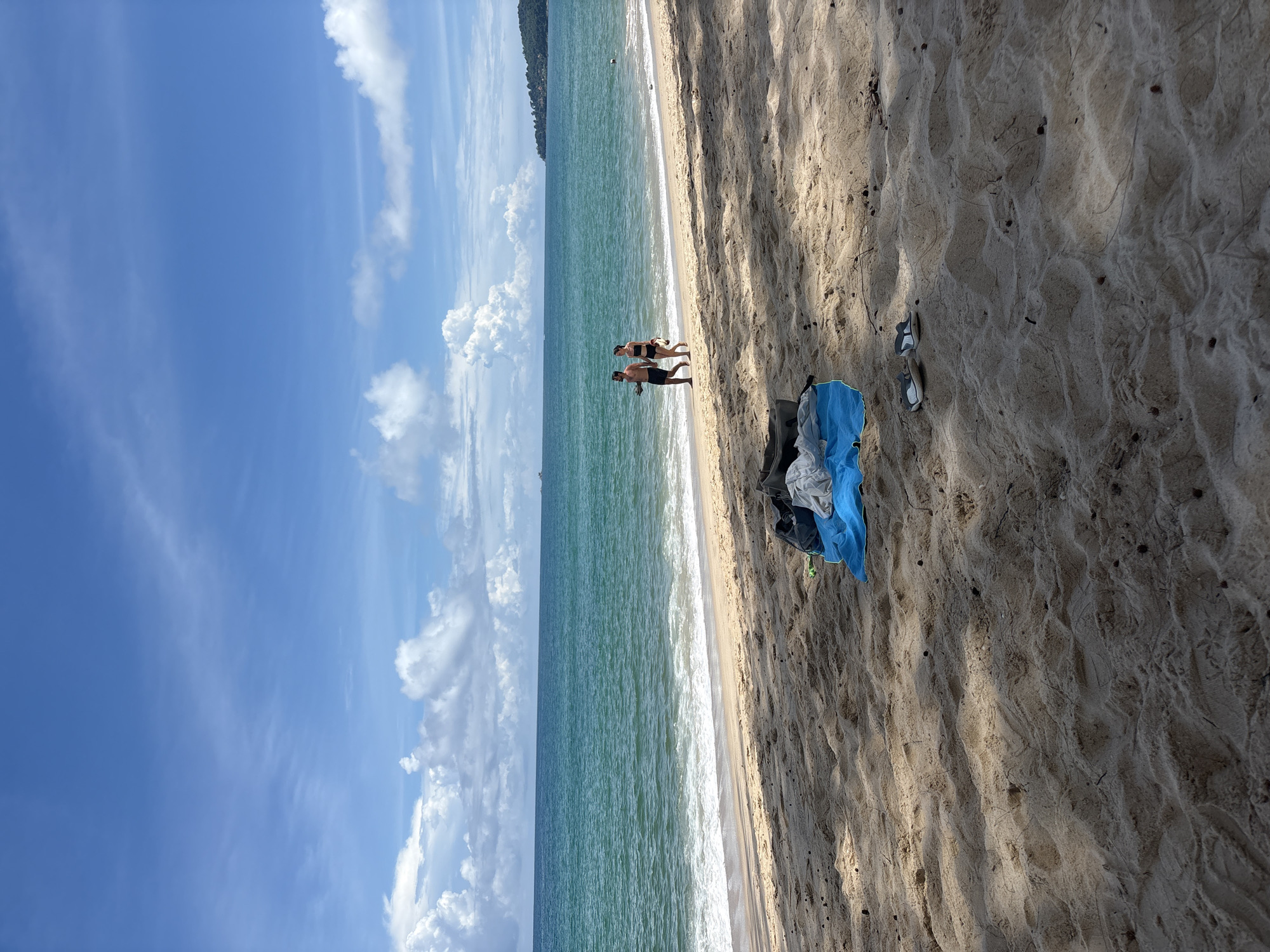 On a sandy beach in Choeng Thale, Thailand, a couple walks near the ocean's edge. The man, wearing dark shorts, and the woman, in a black bikini, stroll hand-in-hand along the wet sand where gentle waves break. The water is a vibrant turquoise, transitioning to a deeper blue on the horizon under a sky filled with scattered white clouds. In the foreground, a blue towel and a dark bag lie on the sand, with a pair of sandals placed nearby. The beach is dappled with shadows cast by overhead trees, suggesting it is mid-day. The scene depicts a relaxed moment on a tropical coastline.