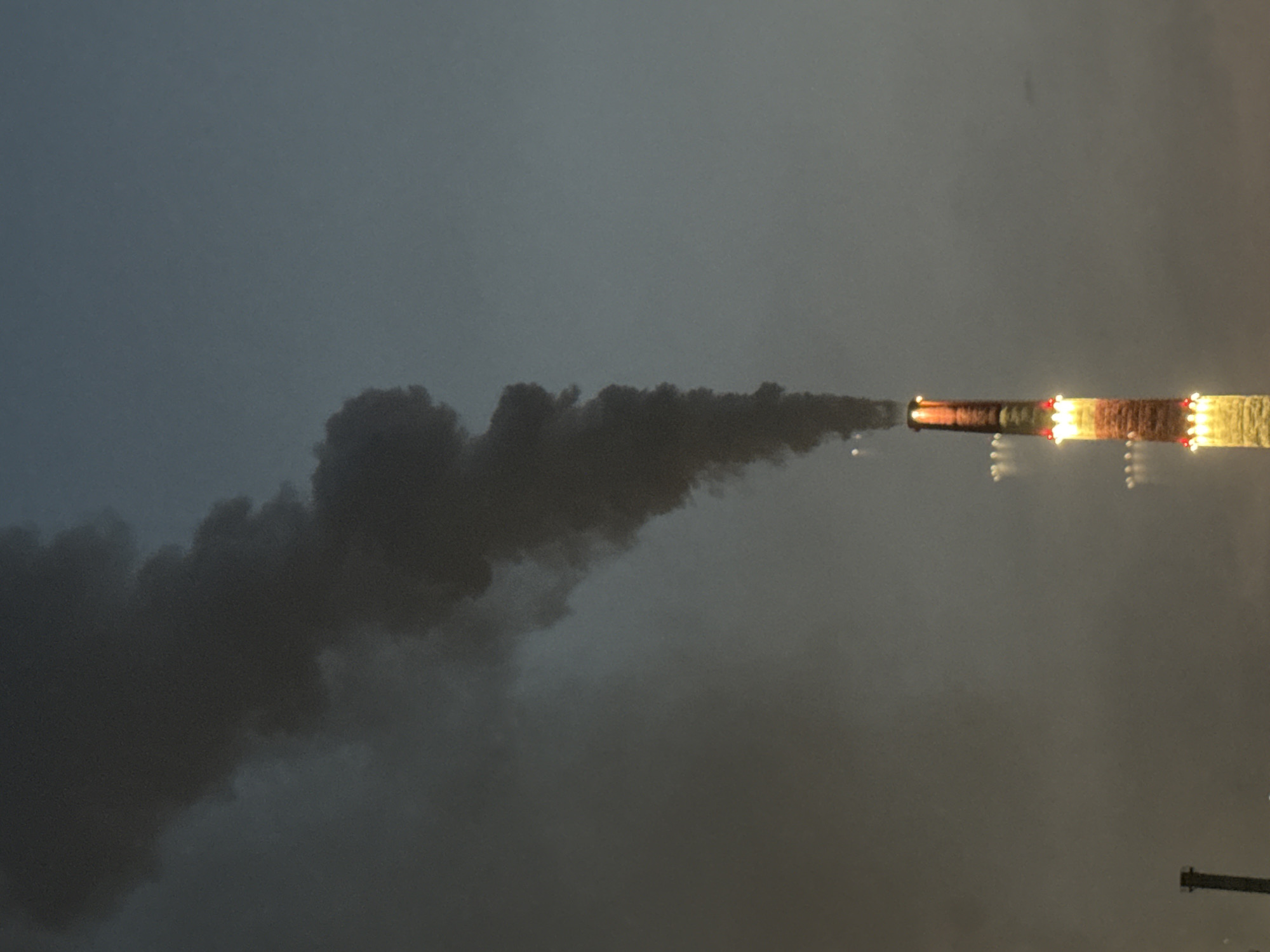 A tall industrial smokestack, illuminated by bands of red and yellow lights, emits a dense plume of dark gray smoke into the overcast sky above Norilsk, Russia. The smokestack, with its striped pattern, stands prominently against the muted blue-gray atmosphere, suggesting twilight or dawn. Distant lights are visible as blurred orbs in the hazy air, and another smaller, darker structure, possibly another industrial element, is partially visible in the lower left quadrant of the image. The overall scene conveys an industrial, possibly cold, environment.