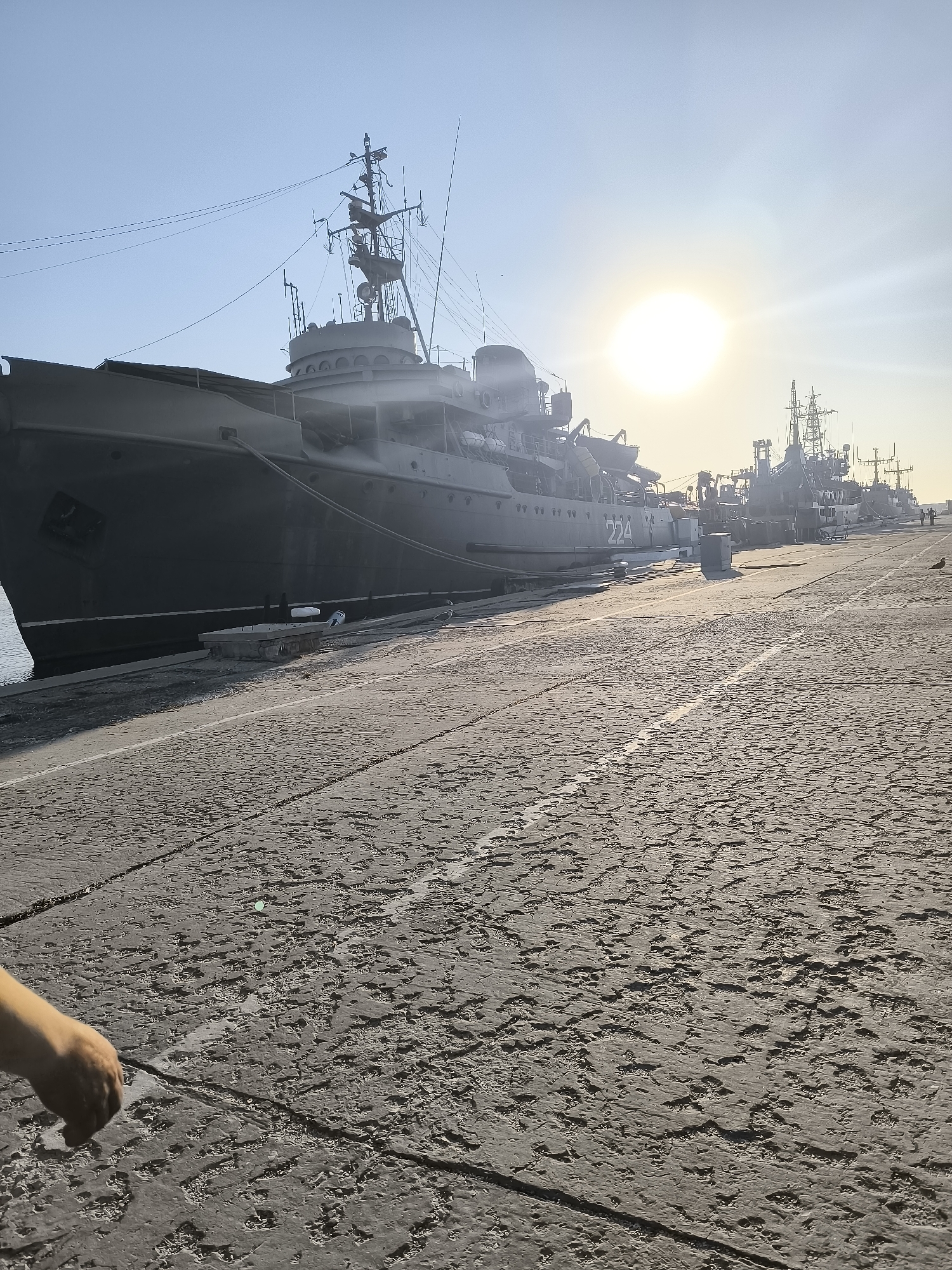 The image depicts a view of the Naval Museum in Varna, Bulgaria. In the foreground, a person's hand is visible, reaching towards the cobblestone pavement. The pavement is weathered and uneven, with visible cracks and textures. The background features a line of naval vessels docked at the harbor. The largest vessel, a dark gray ship with the number "224" visible on its side, is prominently positioned on the left. Other ships, appearing to be various types of warships, are lined up behind it. The sun is visible in the upper right quadrant of the image, creating a bright glare and casting long shadows. The sky is clear and blue. The scene appears to be set during daytime.