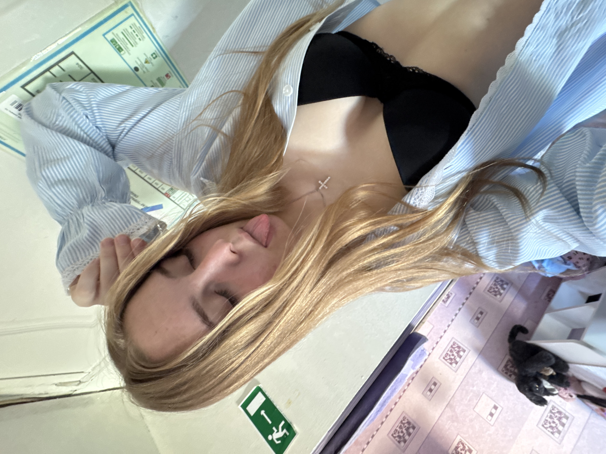 A woman with blonde hair, wearing a light blue striped shirt and a black bra, is lying on a surface. She has her eyes closed and her tongue slightly out. A silver cross necklace is visible on her chest. A black cat is on the floor nearby. The surface the woman is on appears to be a floor or table, with a green and white sign showing an emergency exit. There is also a schematic or diagram on the surface. The floor has a repeating pattern, possibly tiles. Based on the sign, which contains text in Russian, and the location of Perm, it is likely this image was taken in Perm, Russia.