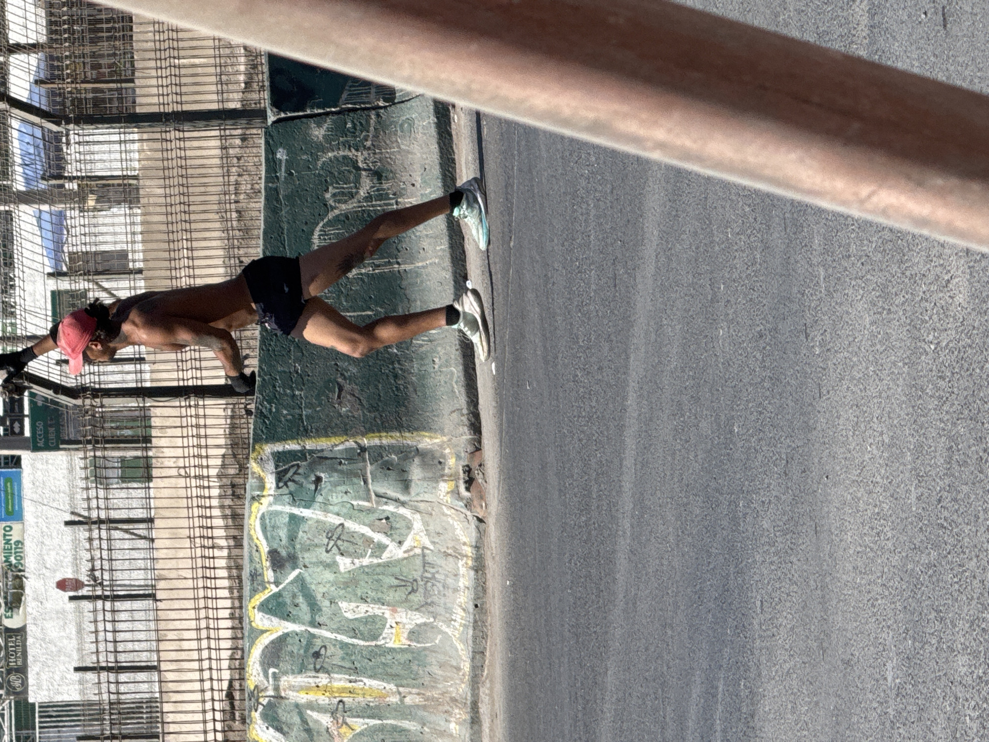 The image displays an urban outdoor scene under bright daylight. A shirtless male figure is captured mid-stride, moving from left to right across the midground. He wears a red baseball cap backward, black athletic shorts, black gloves, black ankle socks, and mismatched athletic shoes (predominantly white/grey on the left foot, light blue/teal on the right). Tattoos are visible on his left arm. The male is positioned adjacent to a dark green concrete barrier, which is heavily covered with white, yellow, and black graffiti. Behind this barrier, a dark metal fence runs horizontally, separating the immediate area from background structures. Visible signs in the background include "HOTEL BENILDA," a partially obscured sign ending in "MIENTO 90119," and a red "PARE" (STOP) sign, indicating a Spanish-speaking region within the city of Unknown, Unknown. The foreground consists of an asphalt road surface, and a blurry, reddish-brown vertical object partially obstructs the right side of the frame.