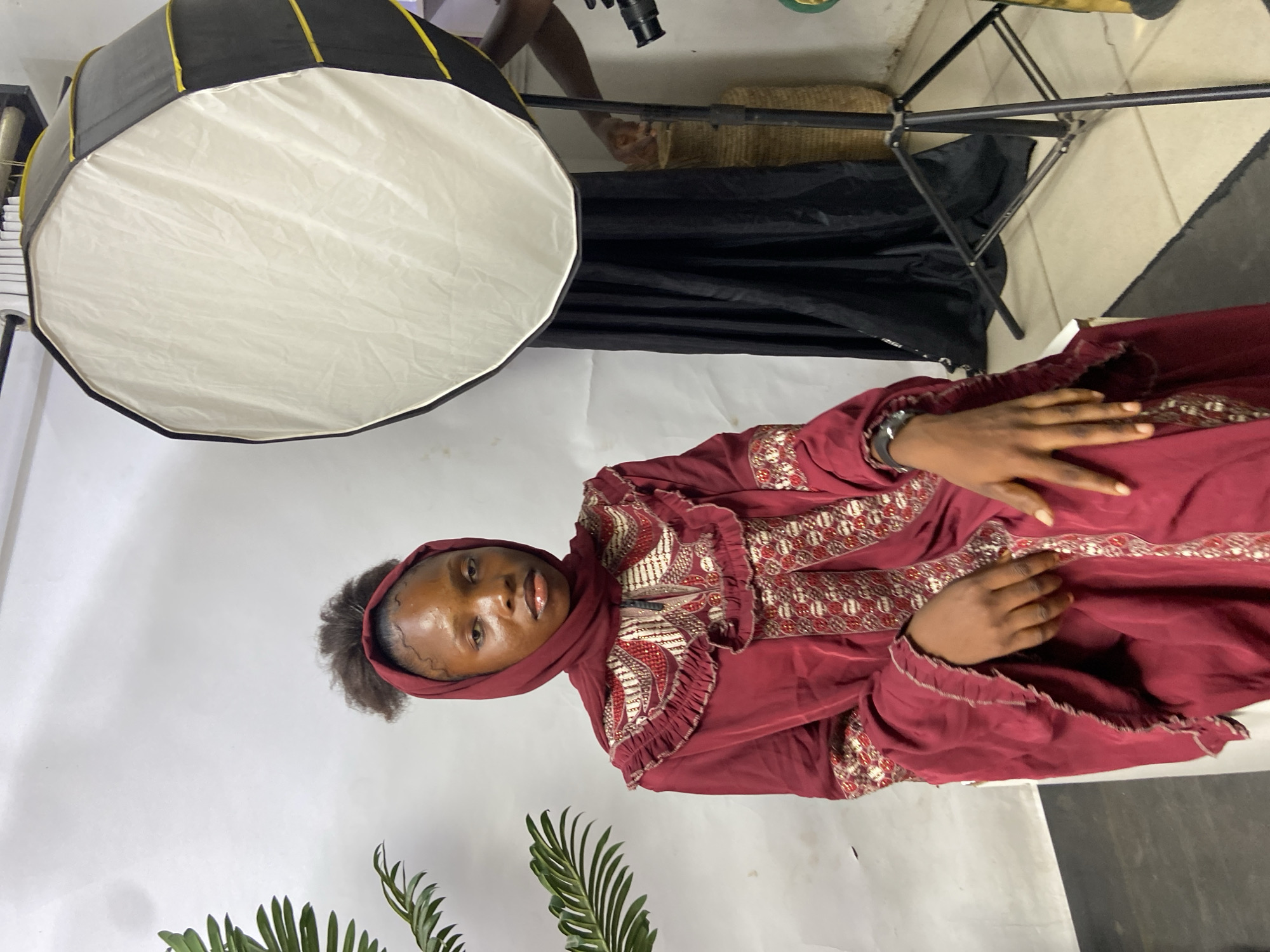 A woman wearing a burgundy dress and headscarf stands in front of a white backdrop. A softbox light is positioned to her left. Part of a fern plant is visible in the lower left corner. The setting appears to be a photography studio. The location is Mararaba, Nigeria.