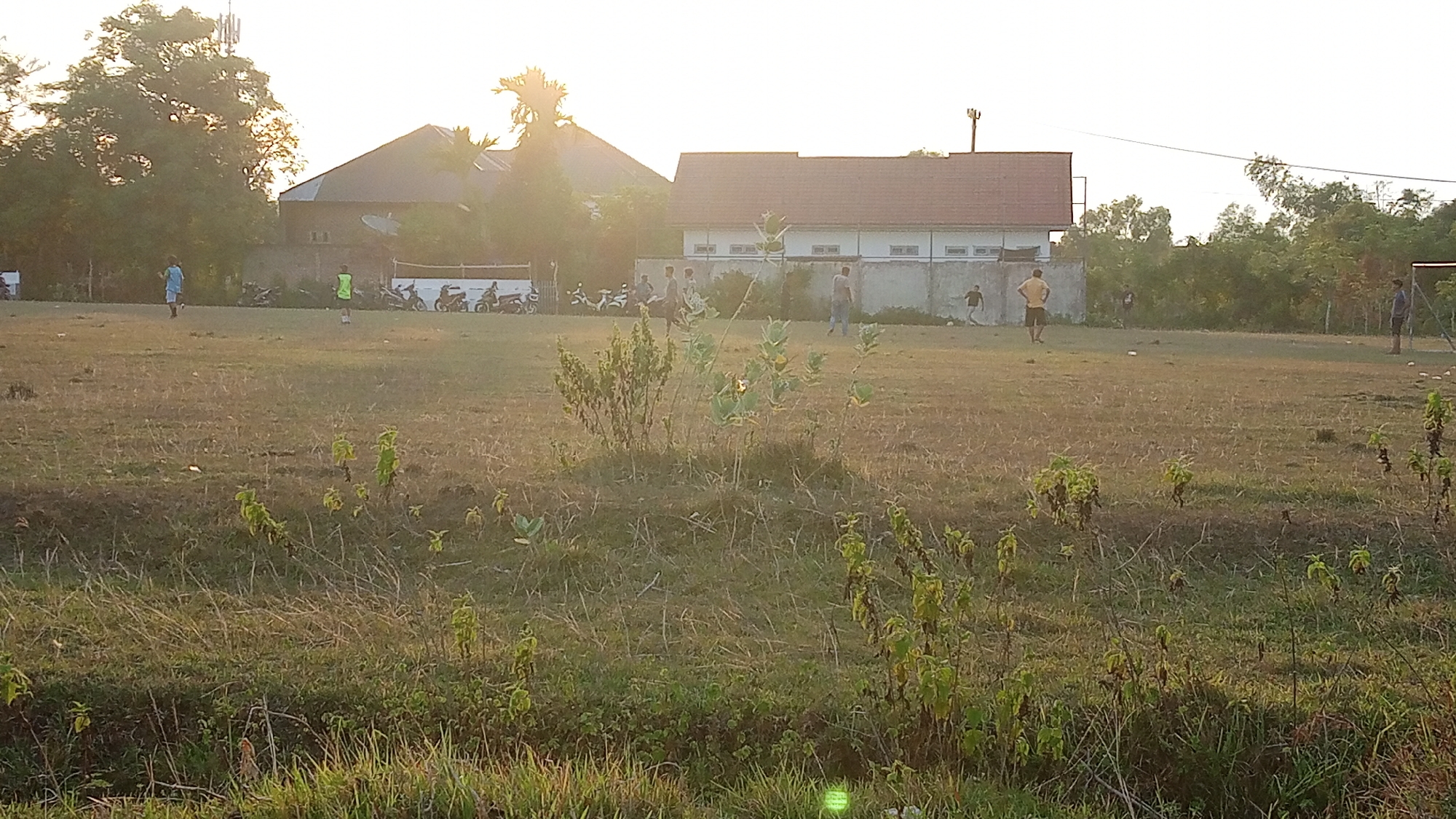 This is a wide, outdoor shot of a soccer field at dusk in Uteunreutoh, Indonesia. The field is mostly dry grass with some patches of green. There are several people scattered across the field, some of whom appear to be playing soccer. In the background, there are some buildings, including a house and a longer building with a red roof. There are also motorcycles parked near the buildings. The sky is bright and hazy, suggesting it's either sunrise or sunset.