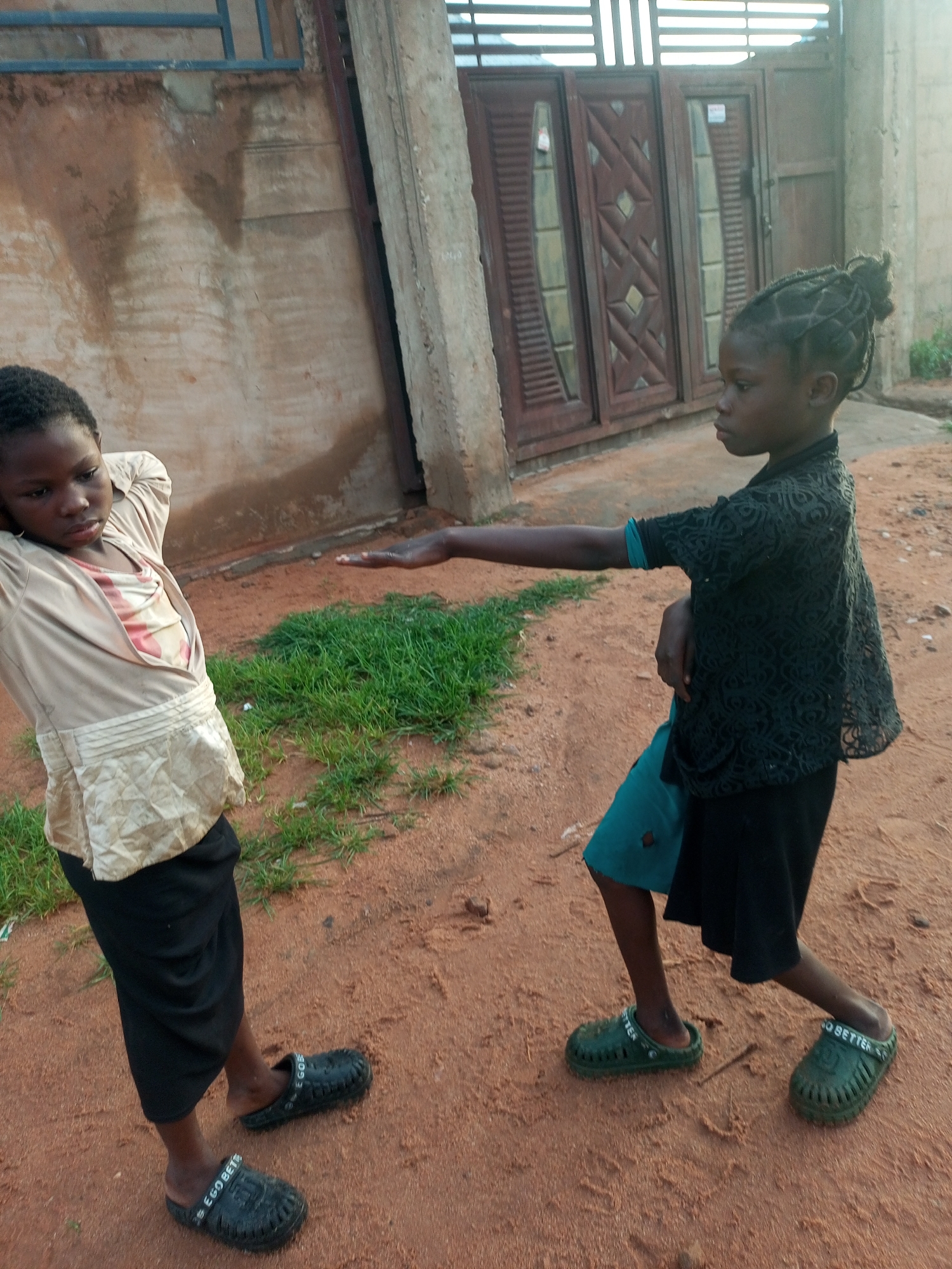 This image captures two young girls in what appears to be an outdoor setting in Tempure, Nigeria. The girl on the left, with her hands behind her head, wears a light-colored, collared shirt layered over a patterned inner garment, a black skirt, and black slip-on shoes. The girl on the right is dressed in a dark, patterned, short-sleeved top, a teal blue undergarment, and a black skirt. She is also wearing green slip-on shoes. Both girls are standing on reddish-brown dirt, with patches of green grass visible.
In the background, there is a weathered building with a large, double wooden door. Above the door, horizontal metal bars form a window or ventilation feature. The building's wall on the left is textured and shows signs of age and possibly water staining.
The activity depicted seems to be a moment of interaction or play between the two girls, with the girl on the right extending her arm. The lighting suggests it is daytime, possibly late afternoon, given the warm tones of the ground and the shadows. The overall scene has a casual, everyday feel. Visible text on the black shoes of the girl on the left reads "EGOBETES". The text on the green shoes of the girl on the right reads "GO BETTER".