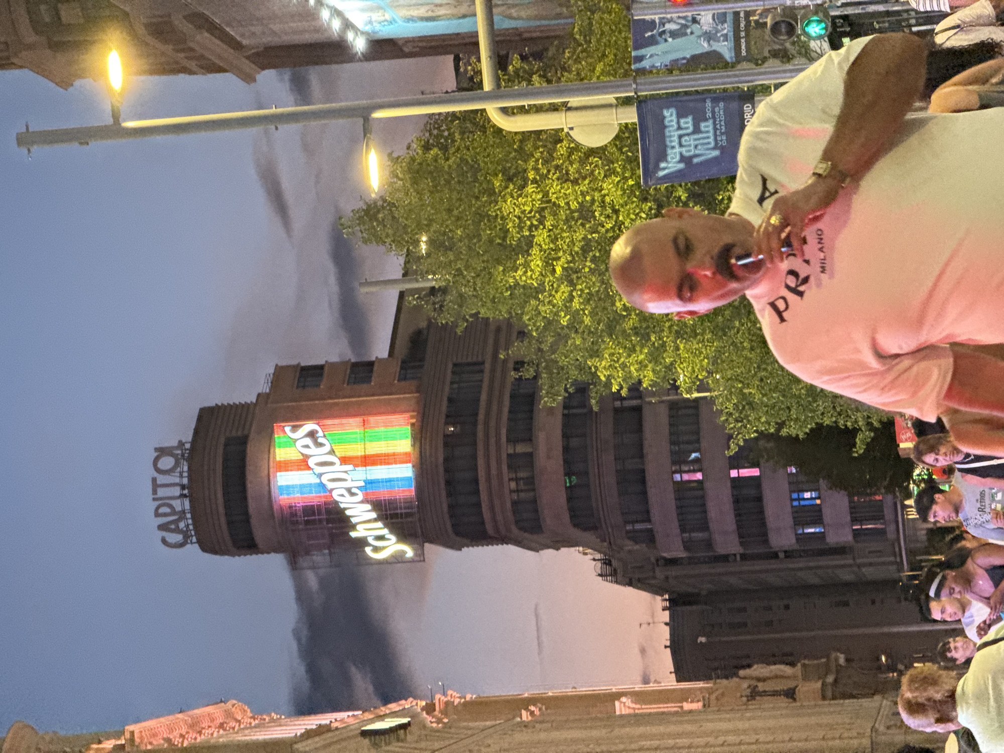 The image captures an evening scene in Madrid, Spain, likely on the Gran Vía, a bustling street known for its architecture and commercial activity. In the background, the iconic Capitol building stands tall, illuminated by a vibrant rainbow-colored "Schweppes" advertisement and the "CAPITOL" sign at its apex. The sky is a dusky blue, with hints of pink from the setting sun.
In the foreground, a man with a shaved head and a prominent mustache is the central subject. He is wearing a white t-shirt with "PRADA MILANO" printed on it and a gold watch on his left wrist. He is holding a vape device to his mouth, appearing to inhale from it. He is surrounded by other people, suggesting a crowded urban environment.
To the right of the man, a lamppost with two illuminated lights casts a warm glow, and a blue banner with text, "Veranos de la Villa 2020 VERANOS DE MADRID," is visible. A green traffic light indicates that traffic may be present or has recently passed through the intersection. The overall atmosphere is lively, characteristic of a city at dusk with active nightlife and pedestrian traffic. The architecture on the left side of the image suggests ornate buildings, common in Madrid's historic center.