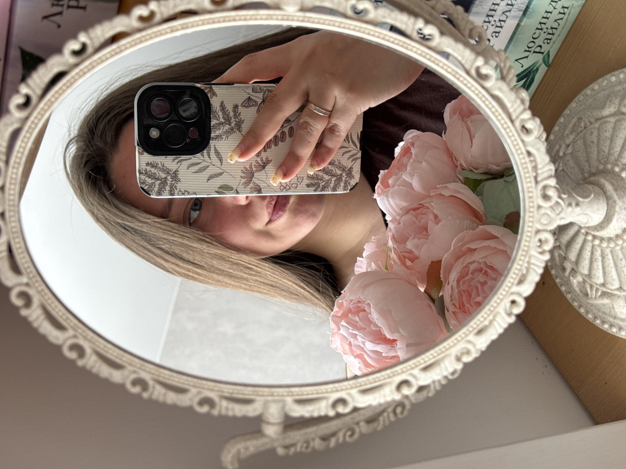 An eye-level shot captures a selfie being taken by a young woman reflected in an ornate, oval-shaped mirror with a cream-colored, decorative frame. The woman's face is partially visible, angled to show her left eye and the lower half of her face, with a neutral to slightly smiling expression. Her blonde or light brown hair frames her face and falls around her shoulders. She holds a smartphone, displaying its patterned case with floral or leafy designs and the triple camera lenses on the back. A simple band-style ring adorns her ring finger, and her nails are painted with a light, natural-looking polish.
To the right within the mirror's reflection, a cluster of artificial pink flowers, resembling peonies, adds a soft, decorative touch. Outside the mirror's reflection, in the blurred background, several objects are visible. A book spine shows discernible Cyrillic text, specifically "ЛЮСИНДА РАЙЛИ," indicating the author Lucinda Riley. Another partially visible word, "син," is above it. Next to the mirror, a white, decorative stand or base with intricate detailing can be seen. The setting appears to be an indoor personal space, possibly a bedroom or vanity area, characterized by soft, ambient lighting. The overall scene conveys a candid moment of self-photography.
