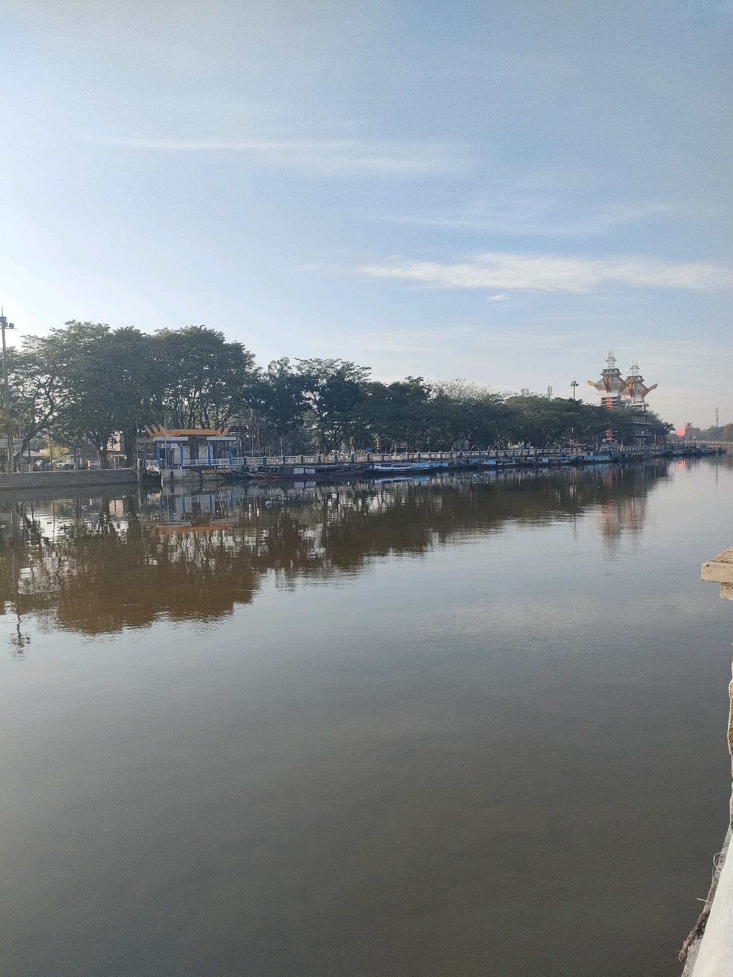 A wide, calm river with a brown tint flows through the city of Banjarmasin, Indonesia. The river reflects the sky and the scene on its banks. On the left bank, lush green trees line a walkway. A small building with a blue roof and yellow accents sits on the embankment, with the words "MENARA PANDANG" visible on its side. Several small boats are docked along the bank. Further down the river on the right bank, a large, multi-story building with a distinctive architectural style, resembling a traditional boat or ship, stands prominently against the pale blue sky. The building has several levels and pointed structures on top. The overall scene suggests a peaceful morning or late afternoon with clear skies and minimal activity.