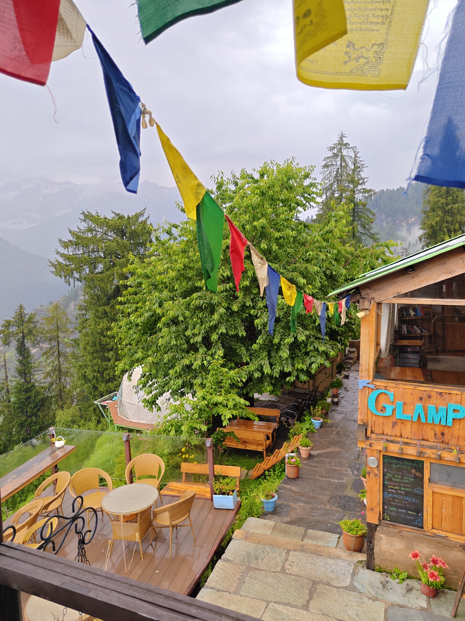 This is an eye-level, medium shot of a glamping site in Prini, India, on a cloudy day. In the foreground, a wooden deck is furnished with four wicker chairs and a wicker table, all wet from rain. The deck is surrounded by a glass railing. Behind the deck, a wooden structure with a sloped roof has the word "GLAMP" in blue lettering. A blackboard menu hangs on the side of the structure. Prayer flags in various colors—red, blue, yellow, green, and white—are strung across the scene. In the background, lush green trees and the misty silhouettes of mountains create a scenic backdrop. A geodesic dome, possibly a glamping tent, is visible on a platform nestled among the trees. The ground is paved with stone and wet.