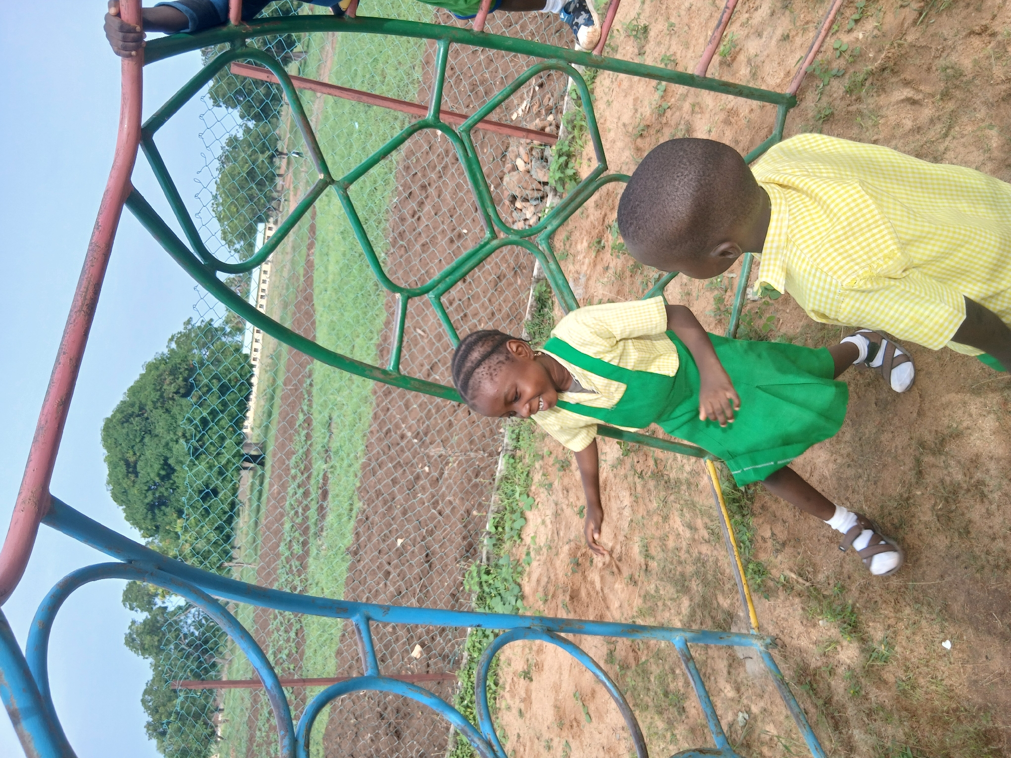 Two children are playing on a climbing frame in what appears to be a playground area. The climbing frame is made of metal bars painted in shades of red, blue and green. A girl is seen in a green and yellow school uniform, is positioned near the center of the frame. A boy wearing a yellow checkered shirt stands to her right. The area around the frame is covered with dirt and grass. A chain-link fence runs parallel to the frame, separating the play area from a ploughed field with distant buildings and trees. This scene likely takes place in Akwanga, Nigeria.