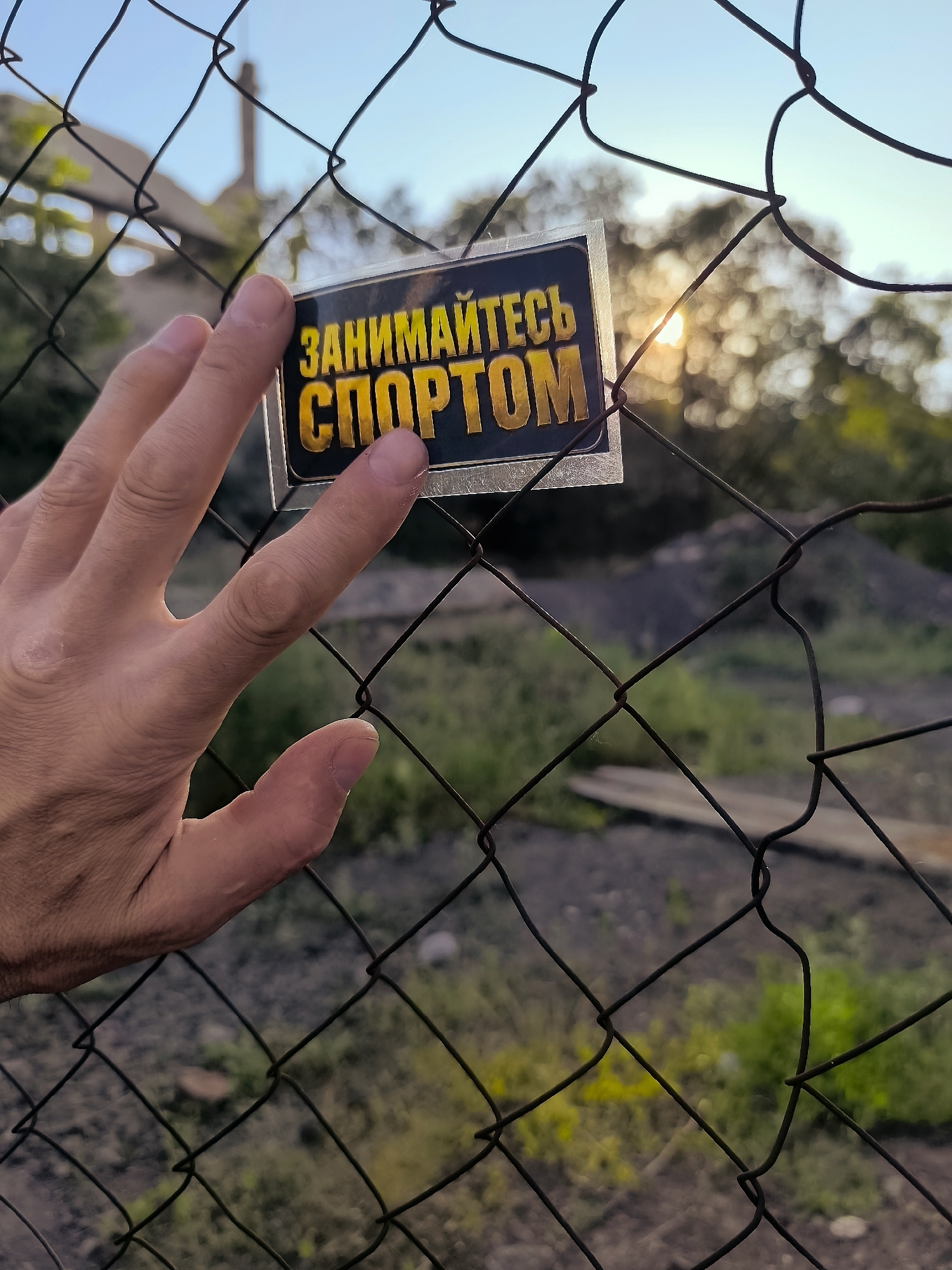 A person's hand is visible in the foreground, with fingers extended and resting on a chain-link fence. Attached to the fence, on the right side of the hand, is a rectangular sticker with yellow text on a dark background. The text appears to be in Cyrillic script and reads "ЗАНИМАЙТЕСЬ СПОРТОМ," which translates to "Engage in Sports." The background is blurred, showing trees, a hazy sunset with the sun visible through the foliage, and a pile of dark material, possibly coal or dirt. The setting appears to be outdoors, with a building or structure faintly visible in the upper left corner. The image is taken at a slightly downward angle, focusing on the hand and the sticker.