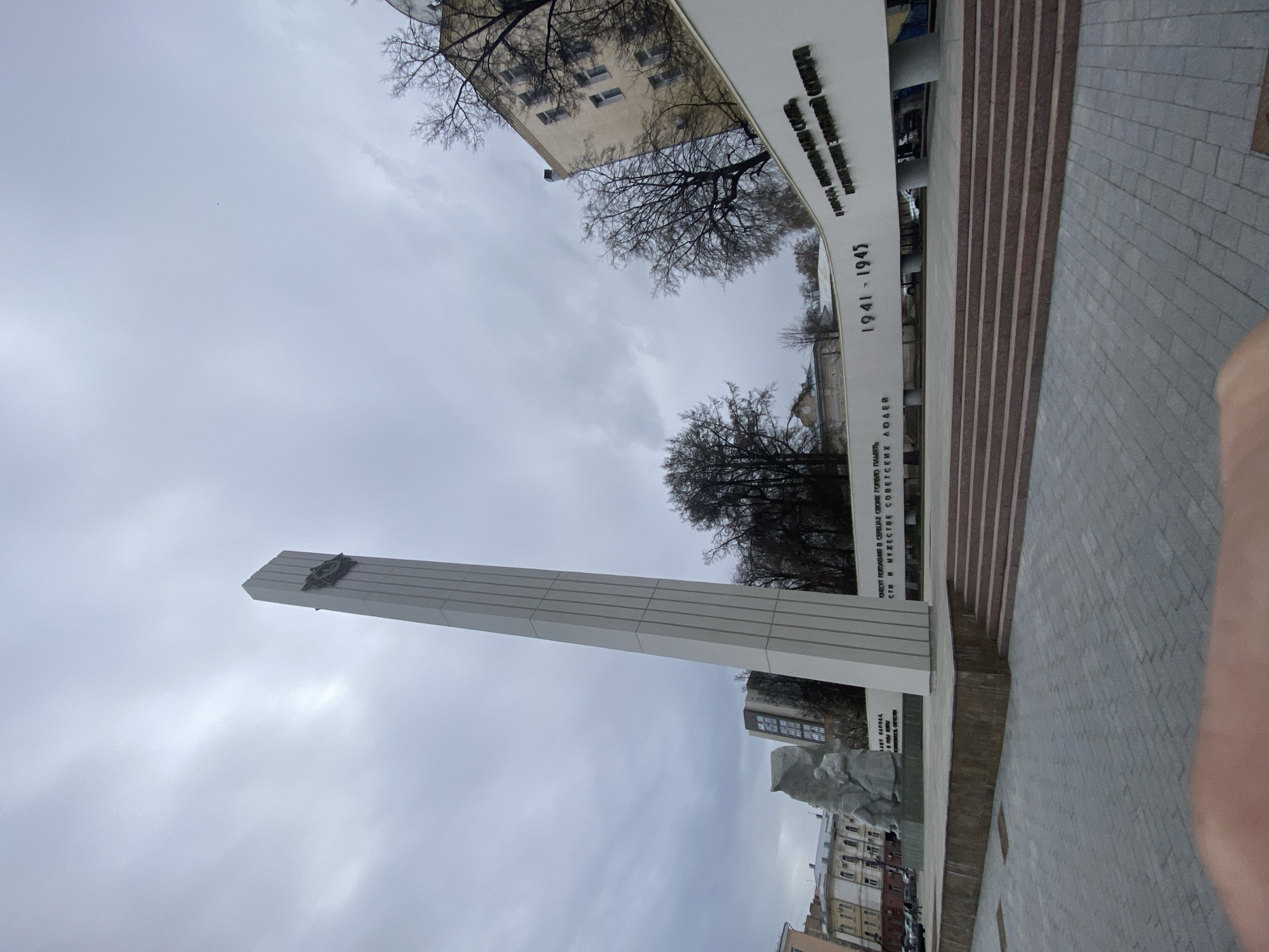 Here is a description of the image:
The image depicts a war memorial in Ryazan, Russia. The memorial is a tall, white obelisk with a symbol at the top, situated in front of a curved, white wall with text in Russian, including the years "1941-1945". A stone sculpture is visible at the base of the obelisk. The environment includes buildings and bare trees in the background. The sky is overcast. The foreground is a paved area with steps leading up to the memorial. The image is taken from a low angle, looking up at the memorial. The bottom right corner of the image has a part of a human finger.