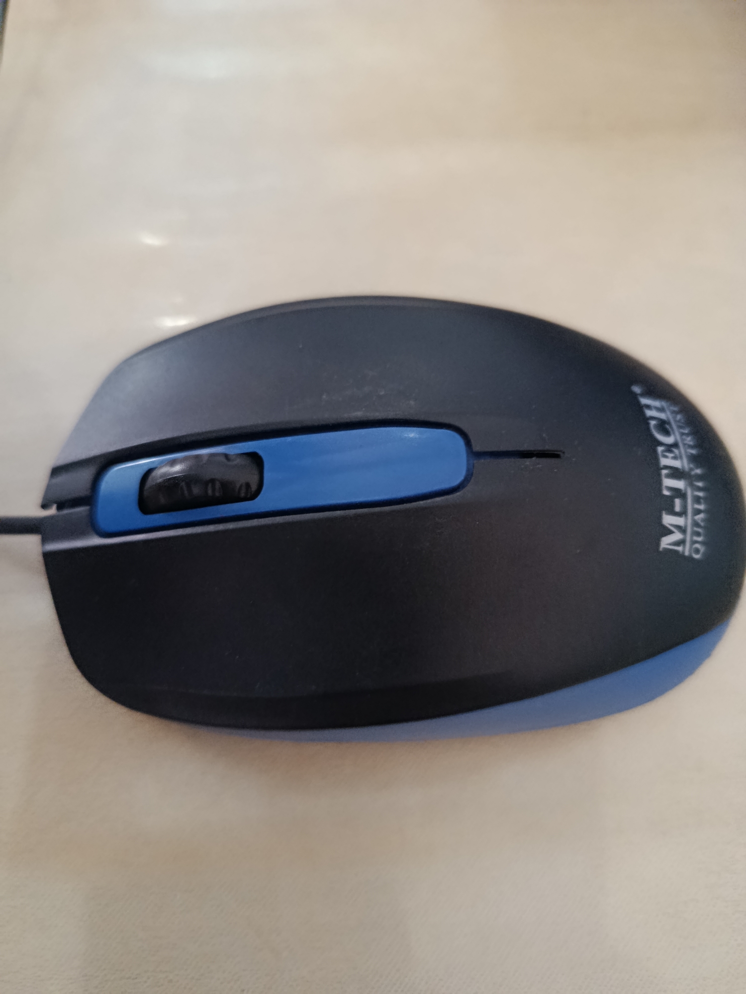 A black and blue computer mouse with the M-TECH logo is placed on a light-colored, textured surface. The mouse has a scroll wheel that is black with visible ridges. The surface appears to be a table or desk. The lighting suggests an indoor environment. The image's perspective is from above, looking down at the mouse. The exact location in Palembang, Indonesia cannot be determined from the image alone.
