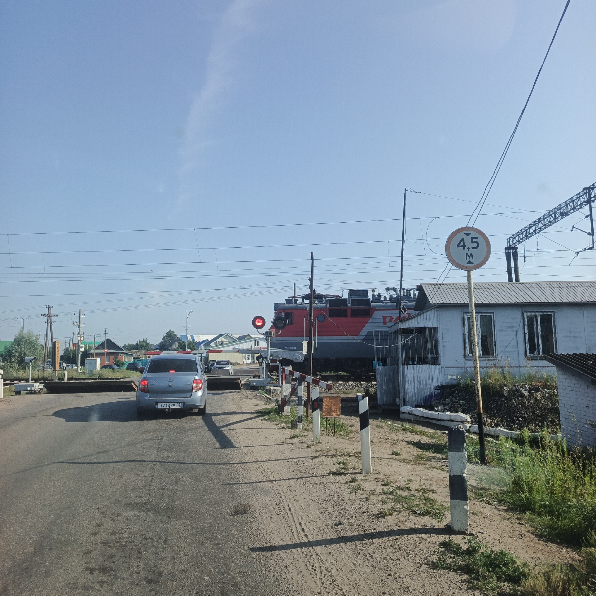 The image depicts a railway crossing in Kambarka, Russia, on a clear, sunny day. A red and grey electric locomotive is stopped at the crossing, with its red signal light illuminated. A blue sedan is stopped behind the locomotive on the road. A circular sign indicating a height limit of 4.5 meters is visible. The crossing is equipped with barriers and striped marker posts. The environment includes a small, light-colored building to the right of the tracks, with vegetation and debris piled nearby. The road surface appears to be asphalt. In the background, there are several houses with pitched roofs, power lines, and trees, suggesting a semi-urban or rural setting.